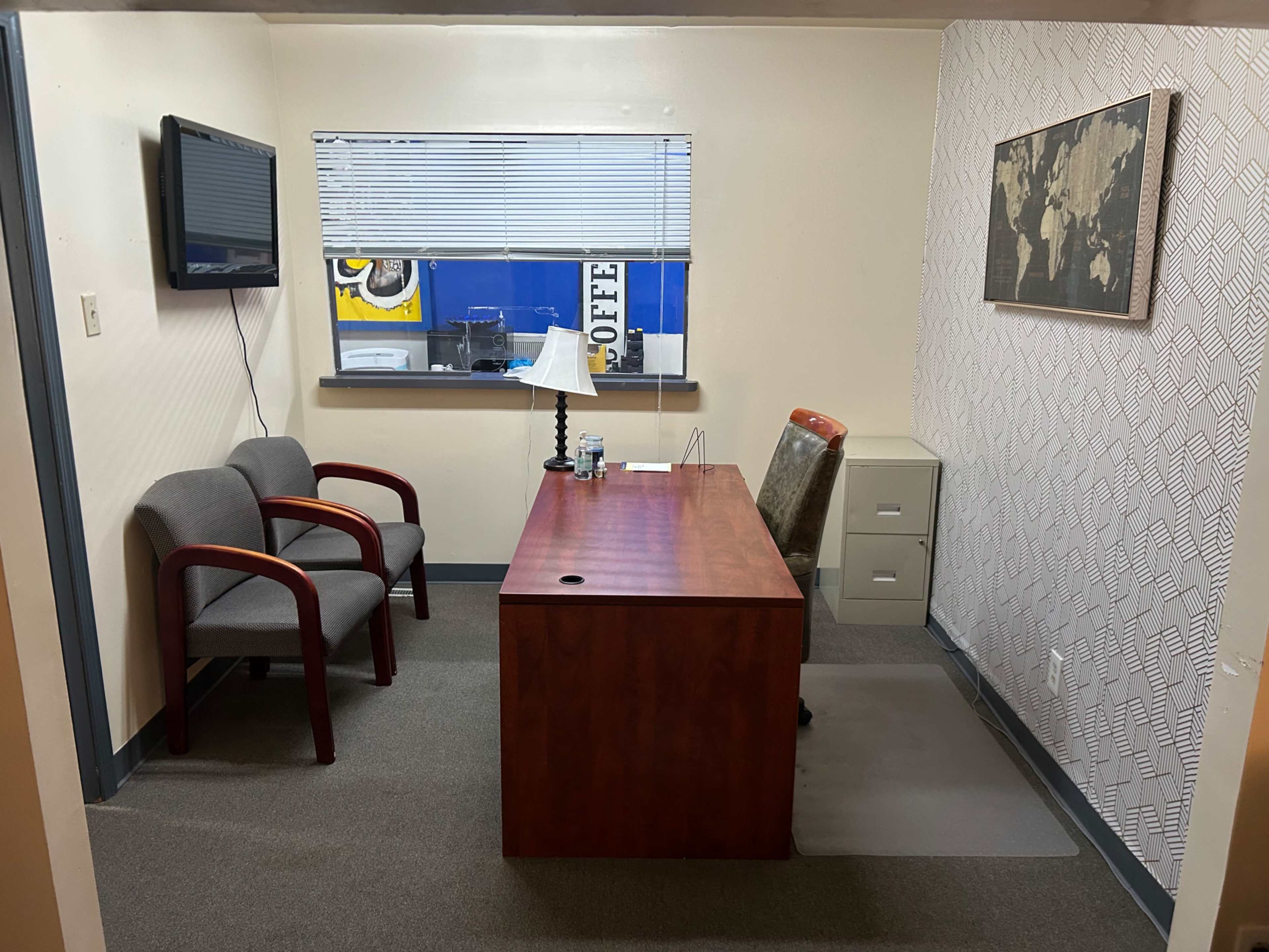The image shows a small office with two chairs, a wooden desk, a filing cabinet, a lamp, and a television mounted on the wall.