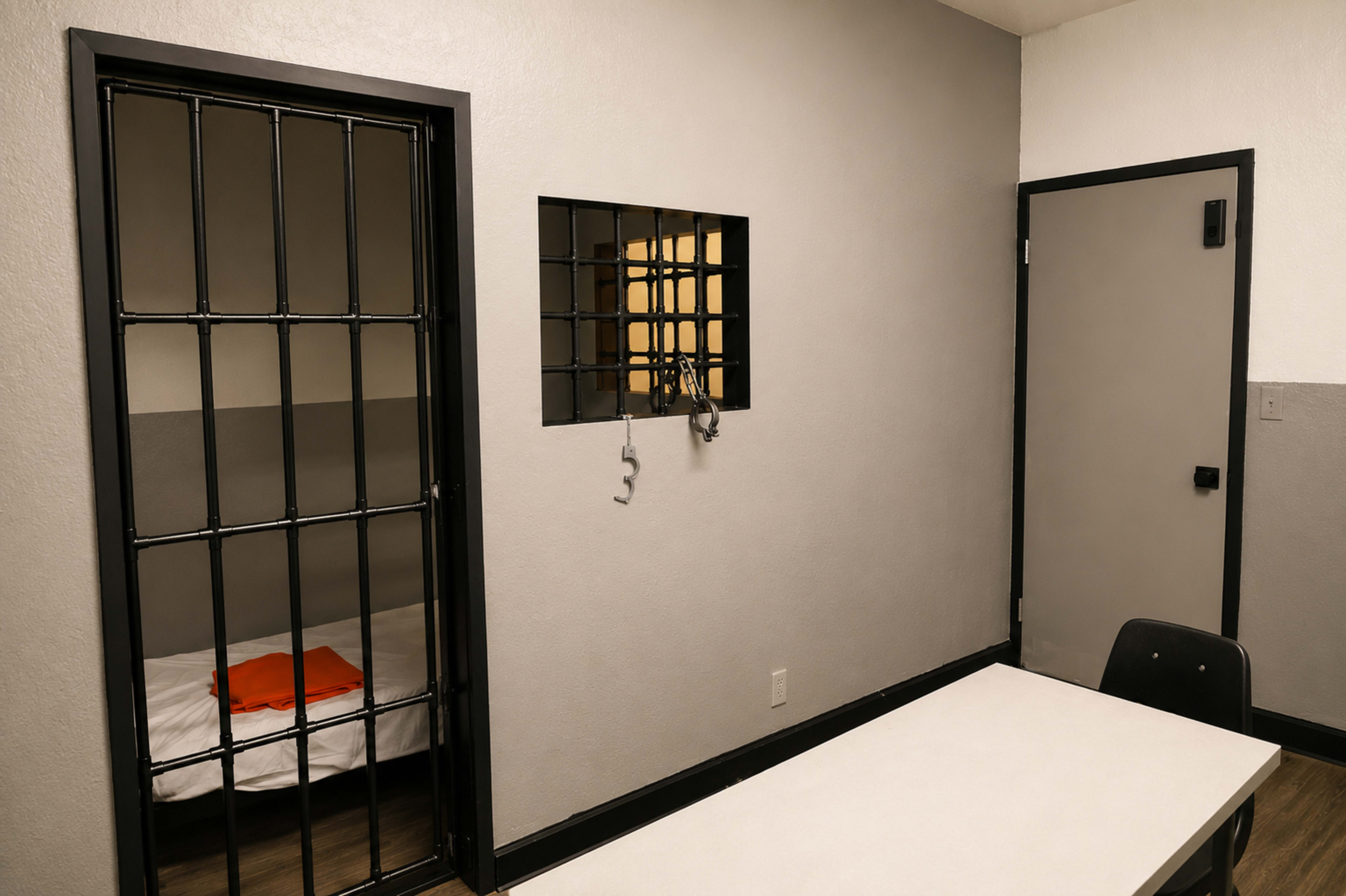 The image shows a stark, small jail cell featuring a barred door, a bare bed with an orange blanket, and a table with a chair in an adjacent area.