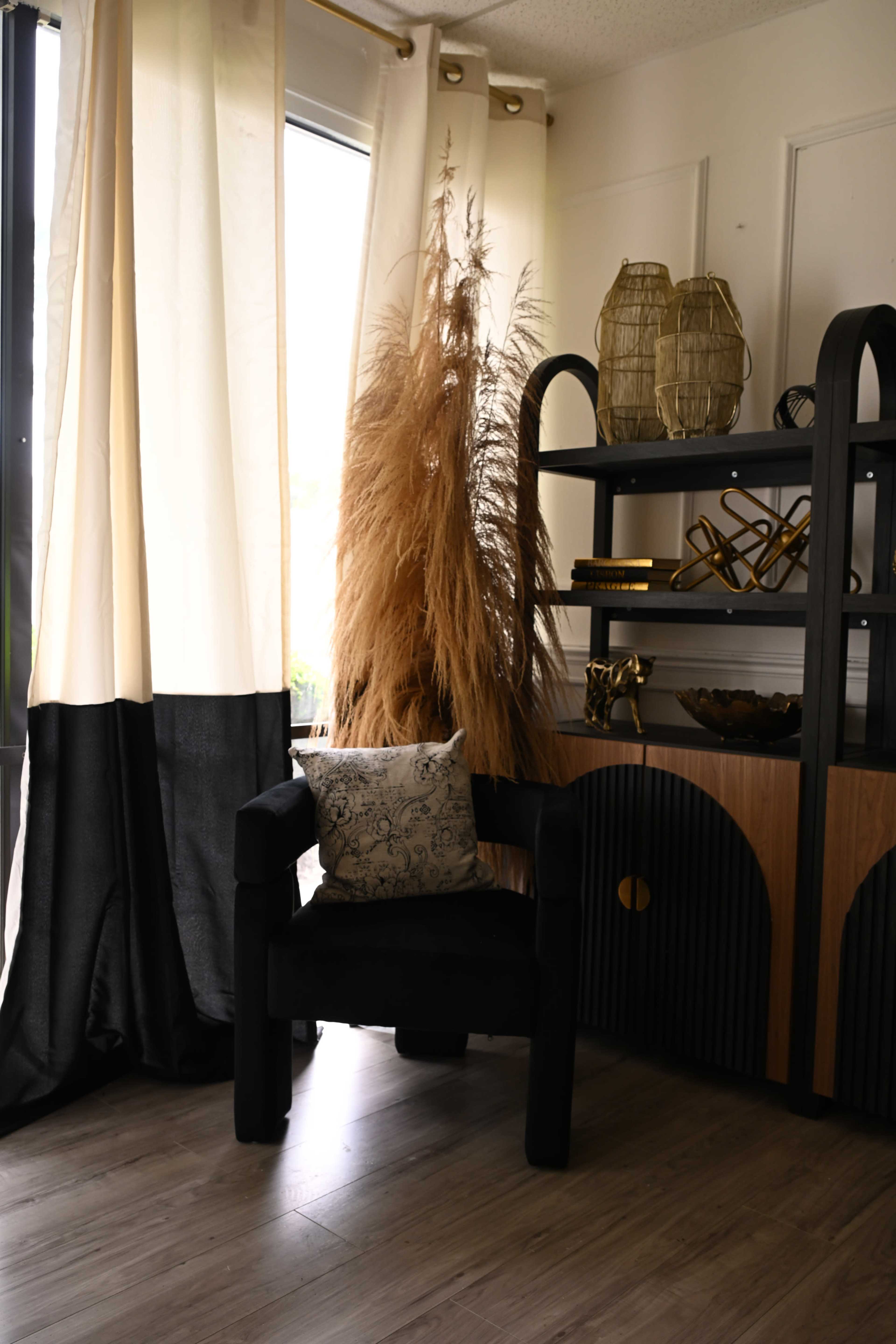 A corner of a room features a black chair with a decorative pillow, a tall dried plant, and a shelving unit with decorative items.