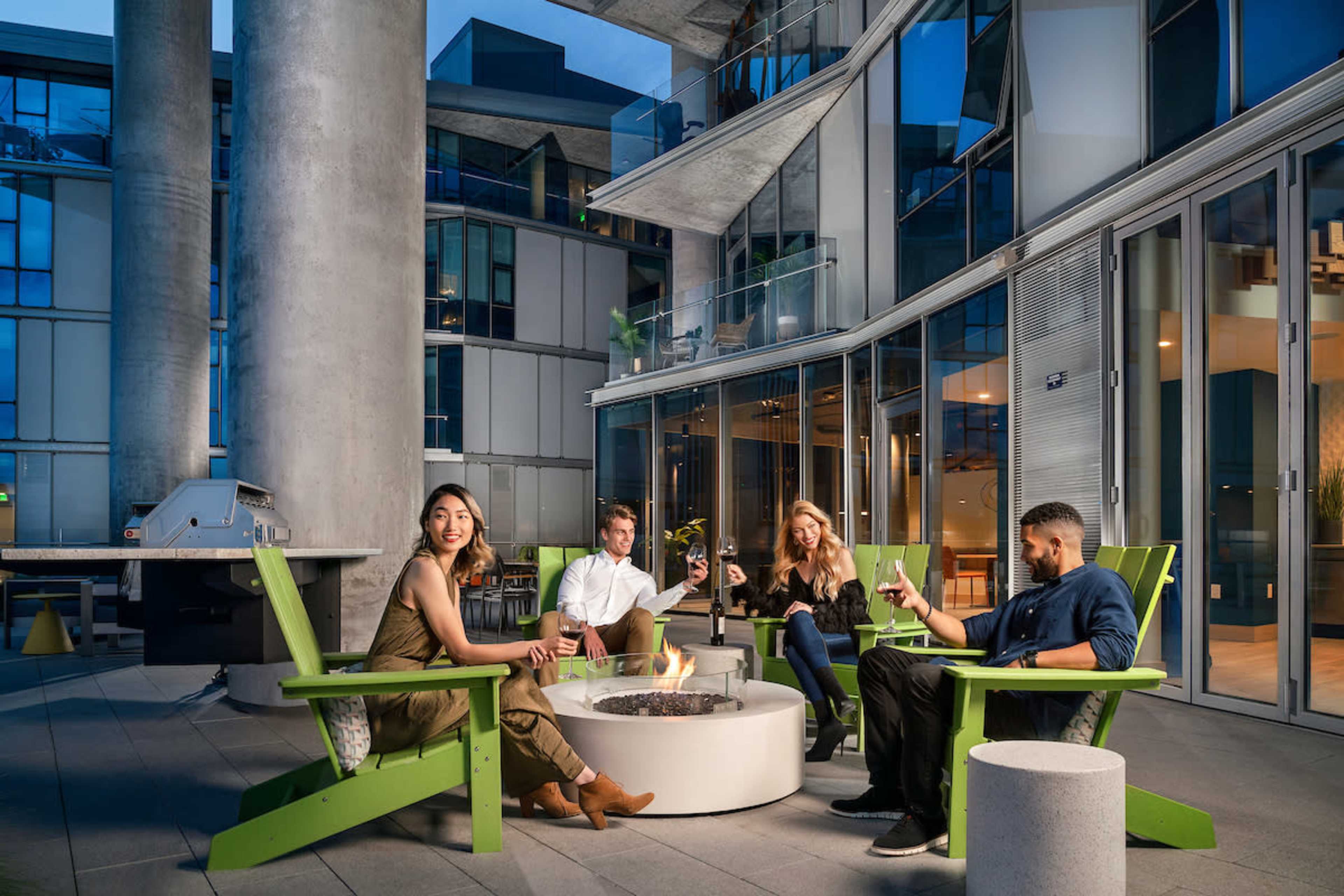 Four people sit around a fire pit in green Adirondack chairs on a modern patio with glass walls and concrete pillars.