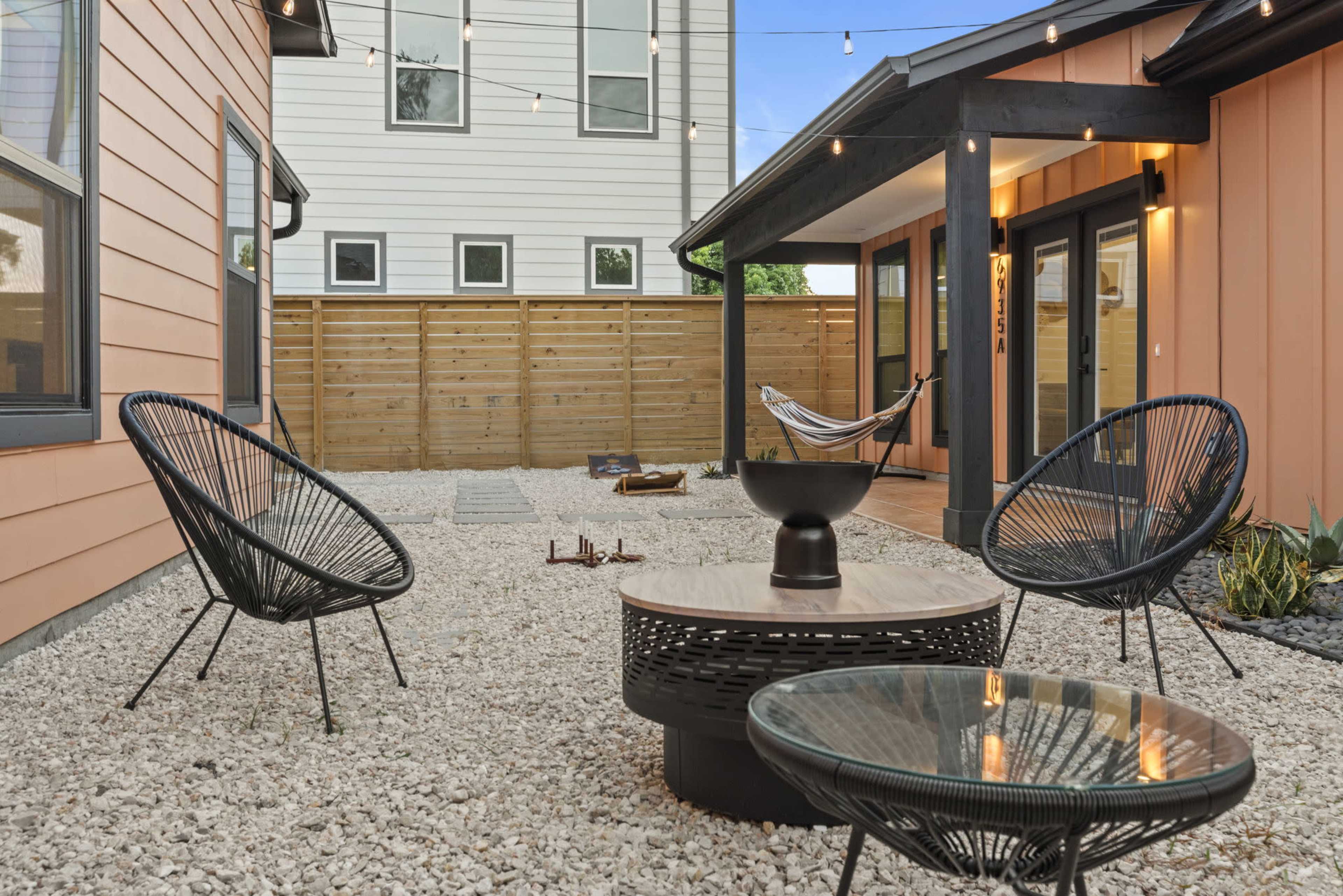 The scene depicts a cozy outdoor patio area with gravel flooring, featuring two black chairs, a central table, a small fire pit, and a hammock nearby, surrounded by wooden fencing and string lights above.