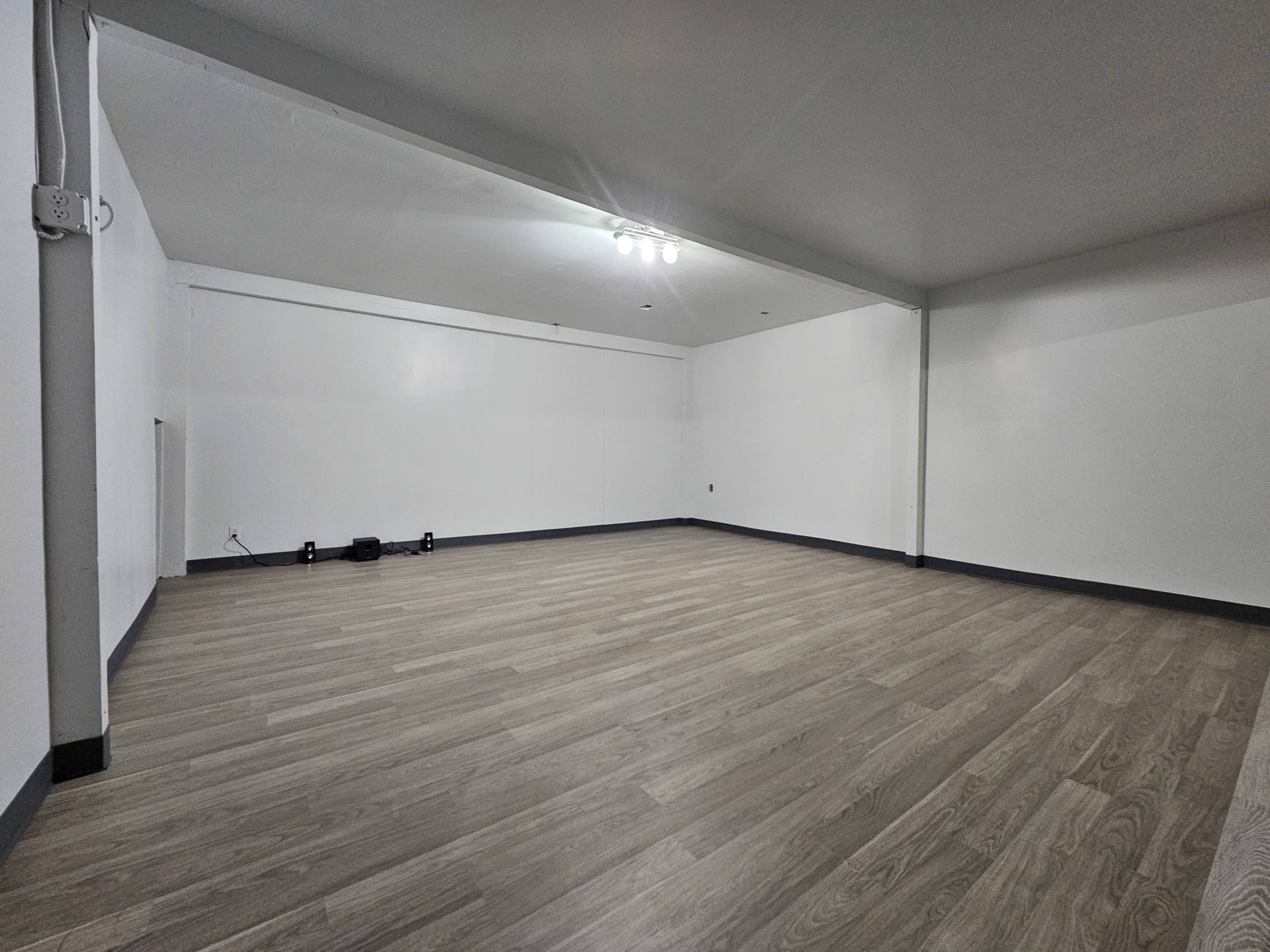 The image shows an empty room with light-colored wooden flooring and white walls, featuring overhead lighting.