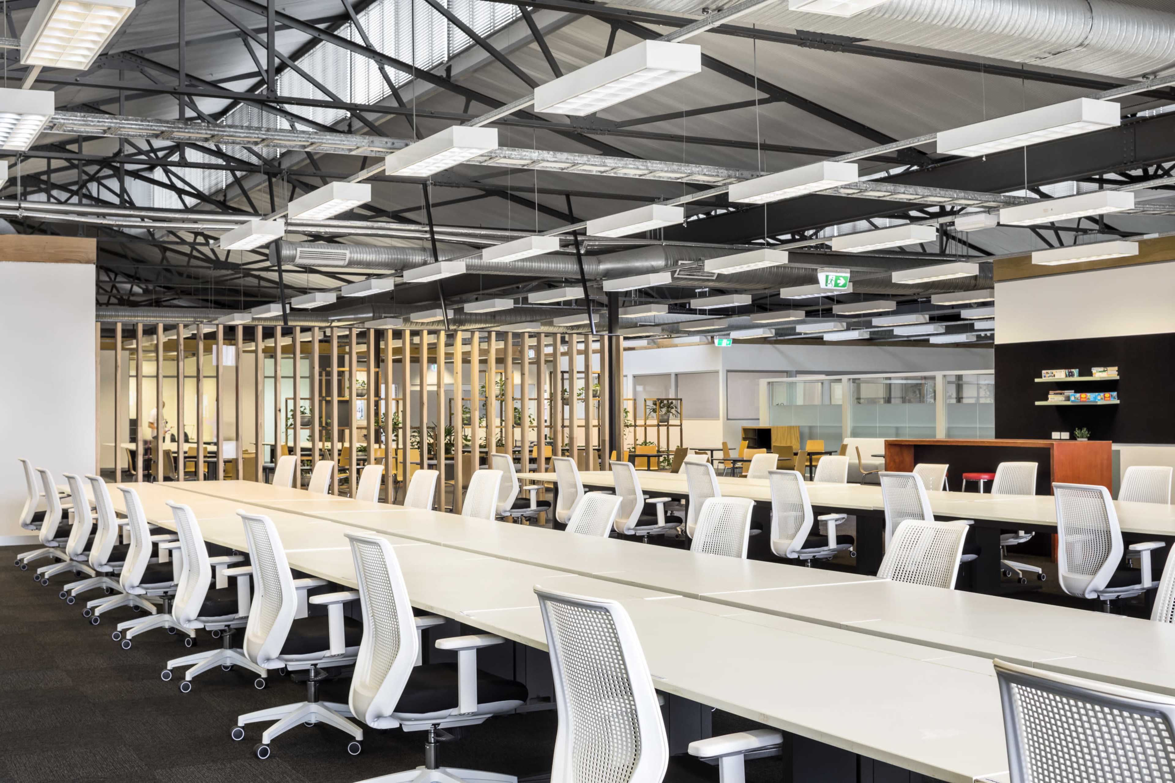 The image shows a spacious, modern office layout with long, white desks and ergonomic chairs arranged in rows, accented by wooden dividers in the background.