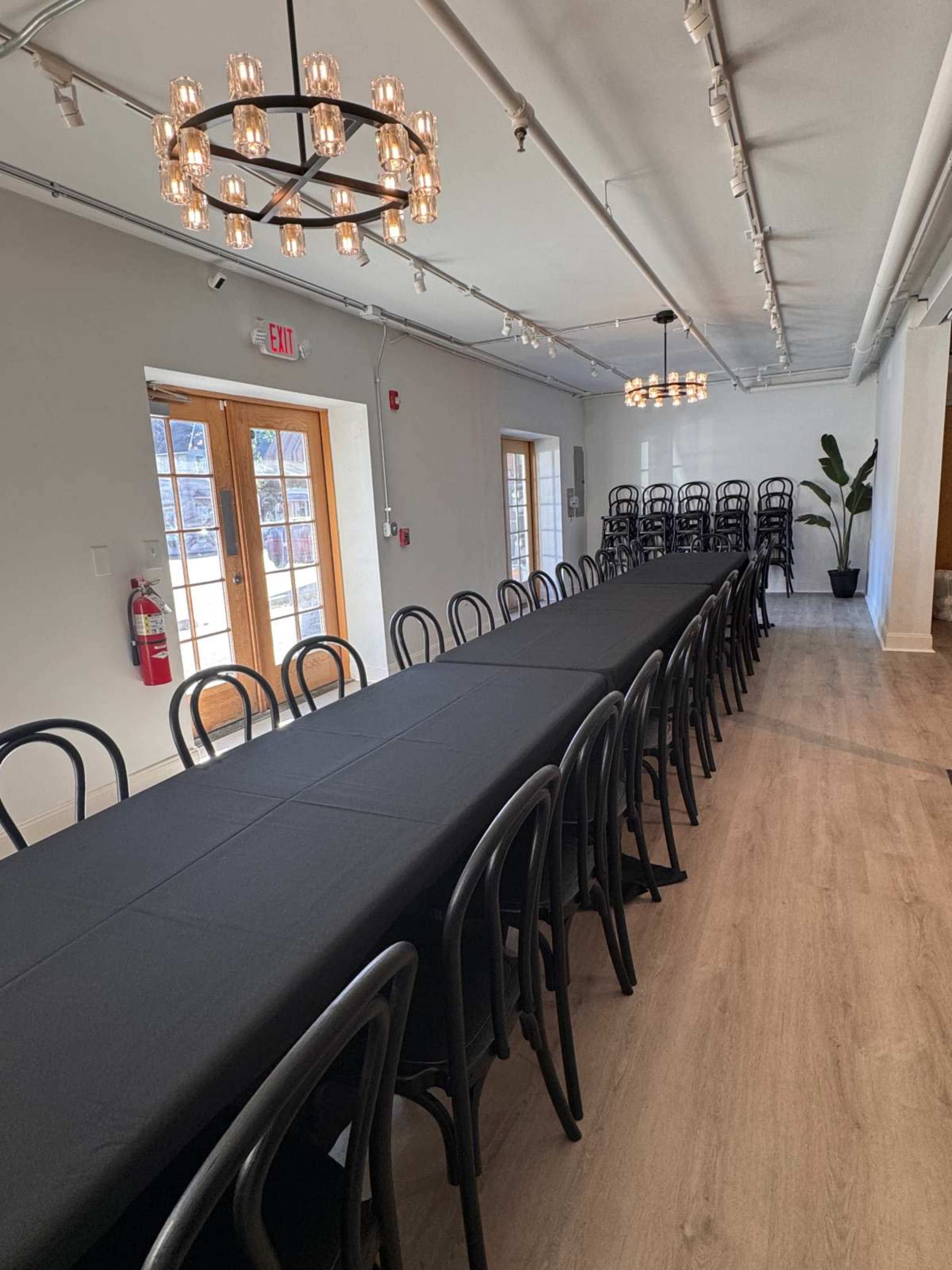 The image shows a long rectangular table set with a black tablecloth in a well-lit room, surrounded by black chairs and featuring decorative lighting fixtures overhead.