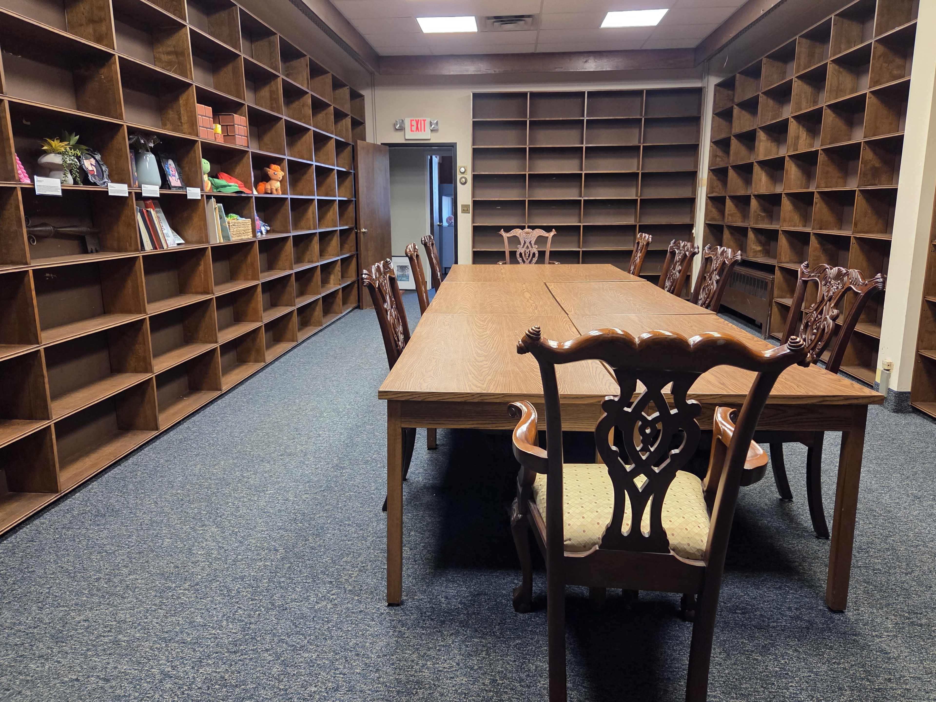 The image shows a spacious room with a large wooden table surrounded by chairs, and empty bookshelves lining the walls.