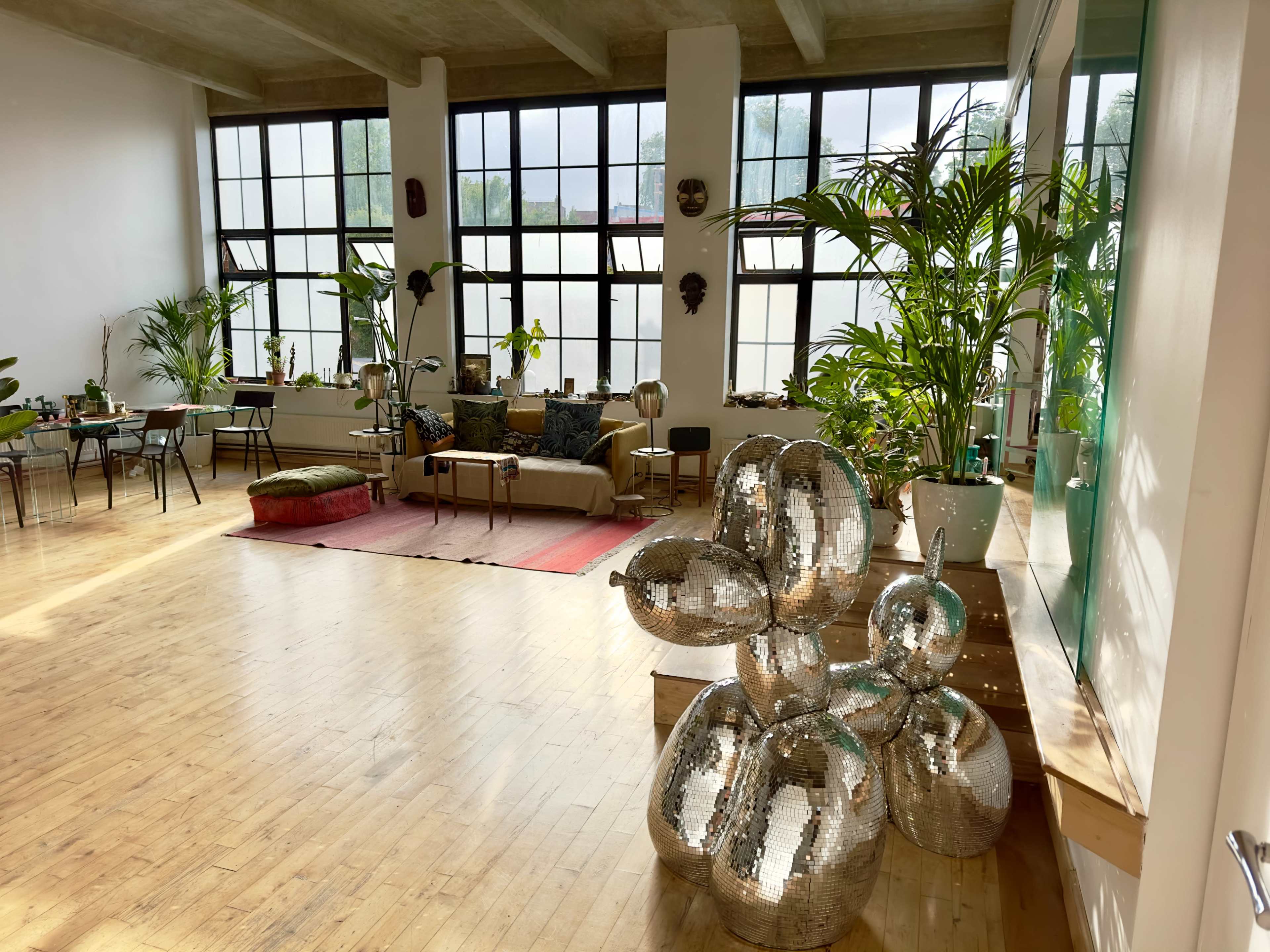 The image shows a bright, spacious room with large windows, wooden floors, various plants, and a reflective sculpture in the foreground.