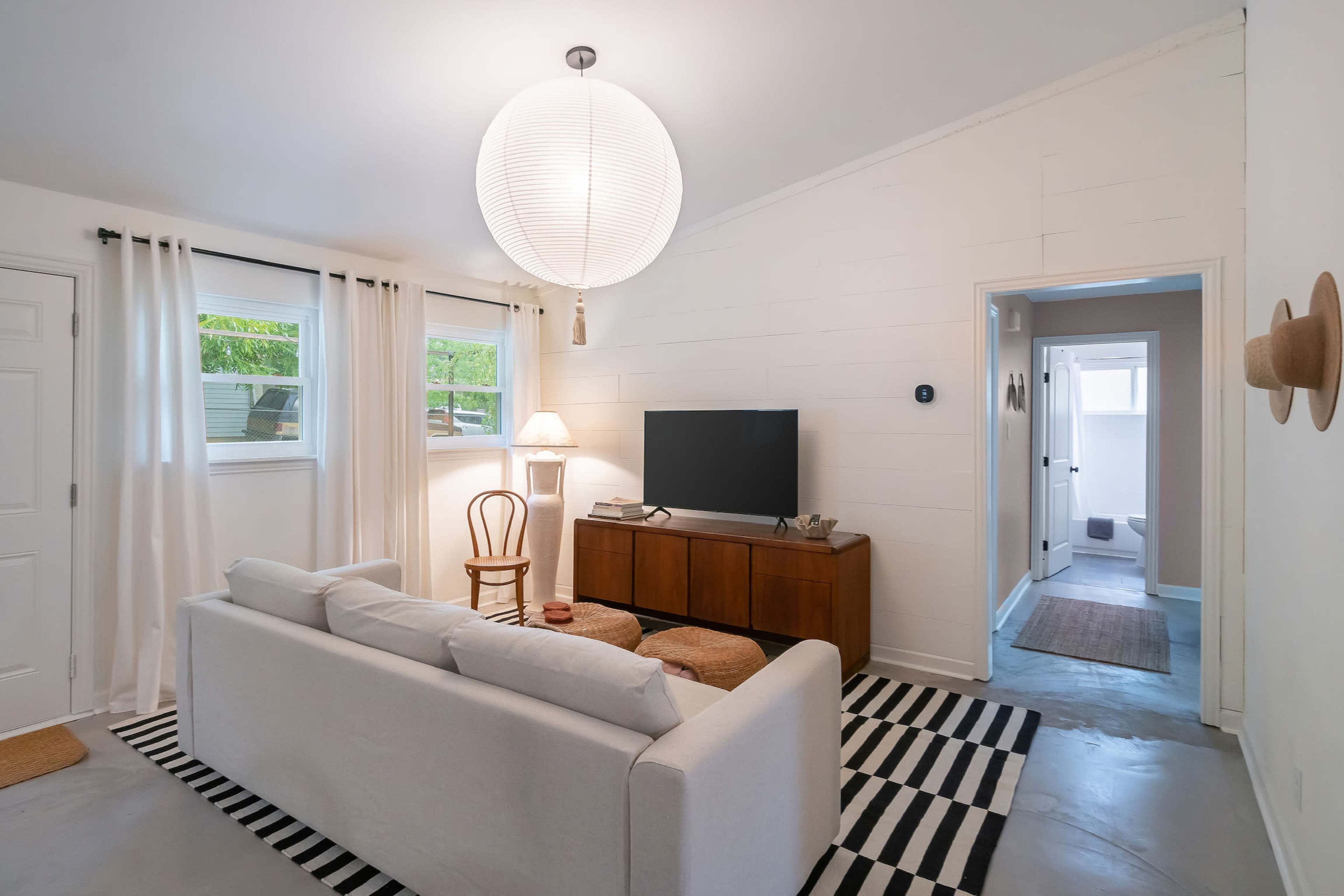 The image shows a bright living room with a white sofa, a wooden TV stand, and a door leading to another room in a modern interior design.
