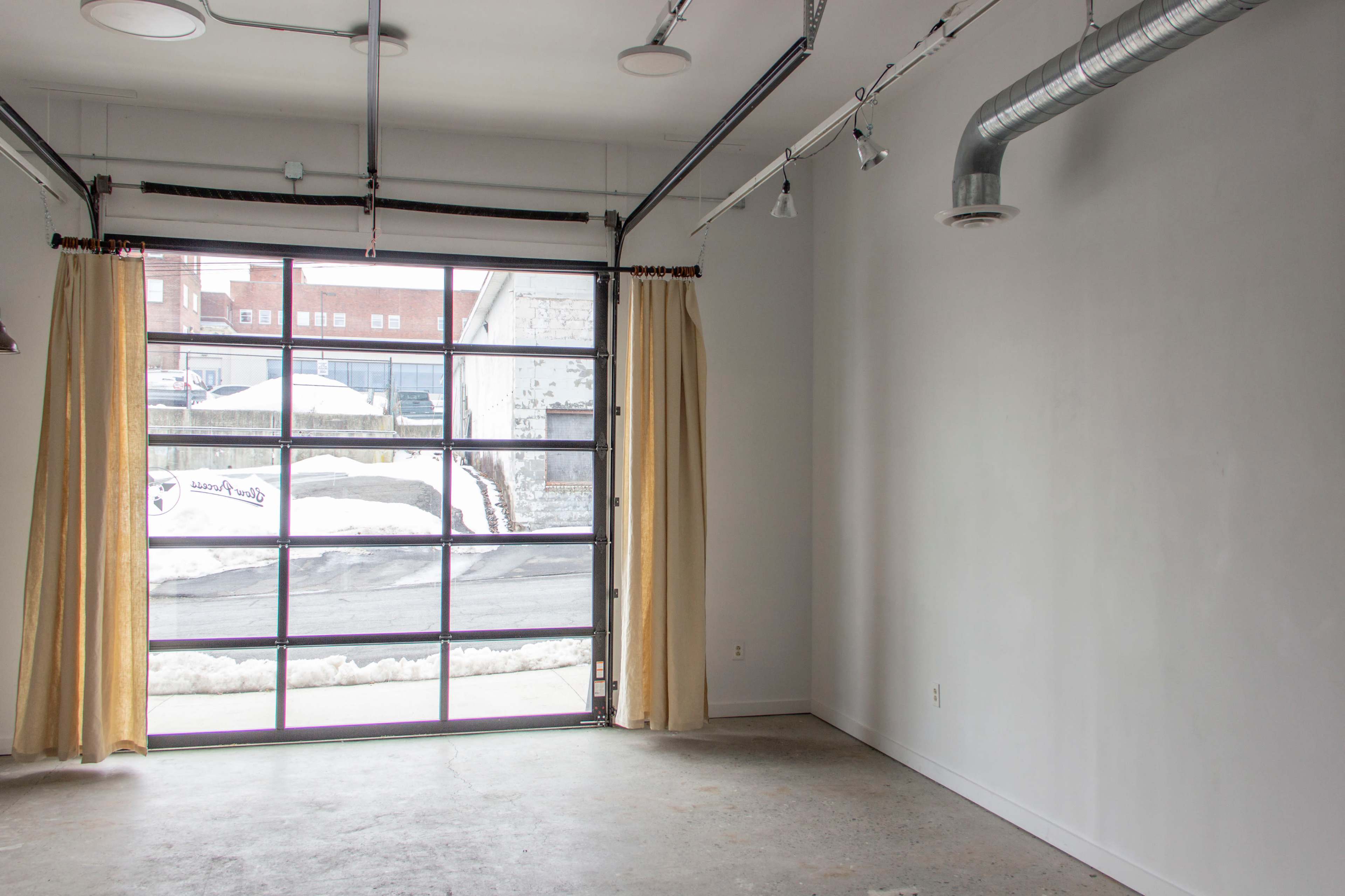 The image shows a vacant room with a large window featuring light-colored curtains, concrete flooring, and exposed pipes on the ceiling.