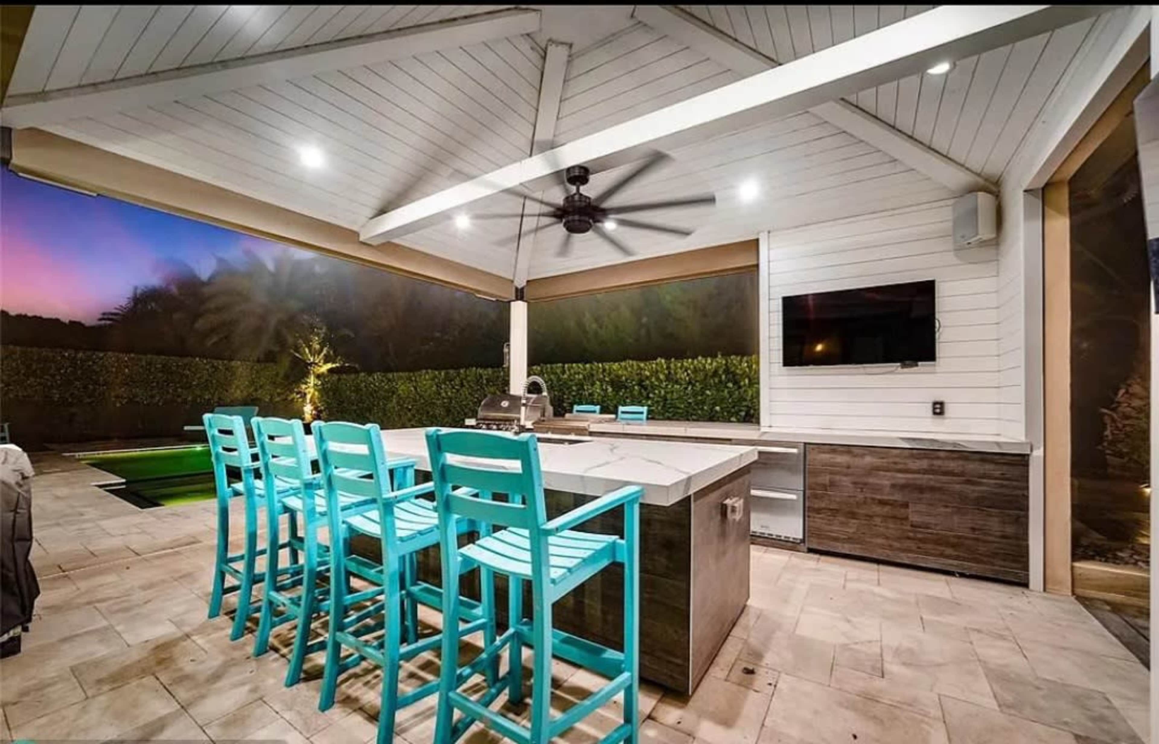 The image shows an outdoor kitchen featuring a bar with turquoise high chairs, a grill, and a television, all under a covered pavilion.