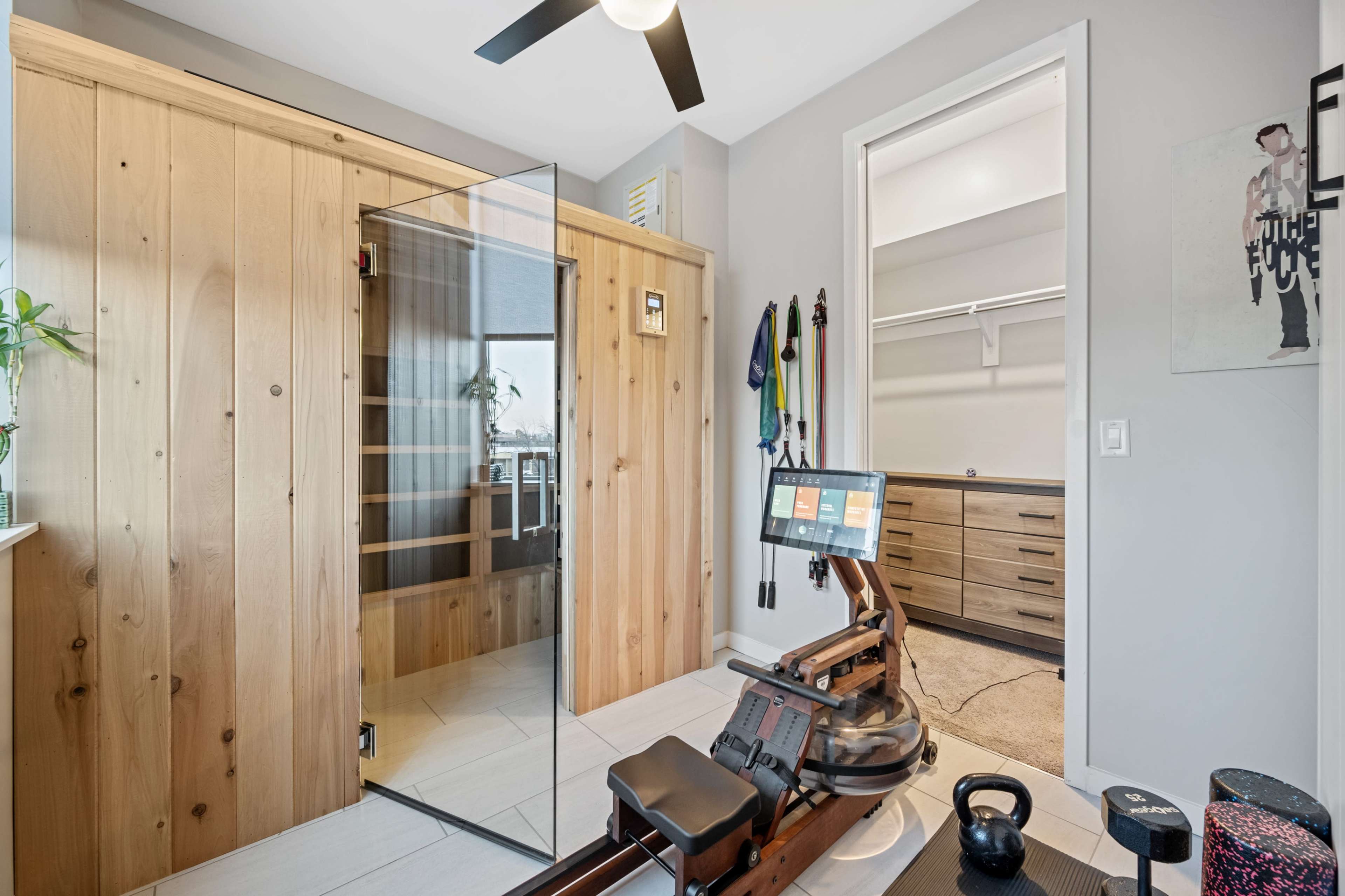The image shows a modern room featuring a wooden sauna with glass doors, a rowing machine, and a closet with sliding doors.