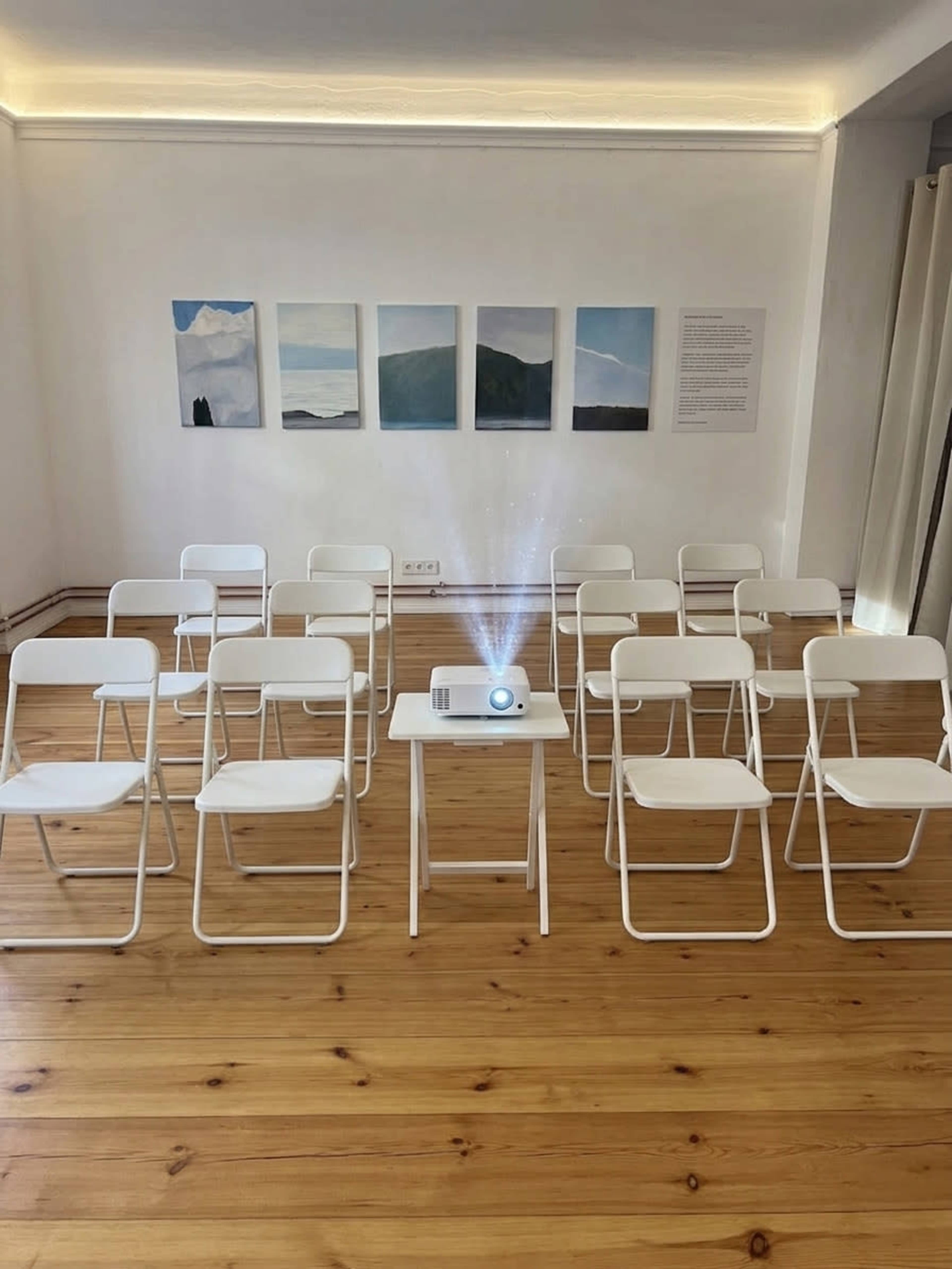 The image shows a sparse room with white folding chairs arranged in rows facing a small table that holds a projector, illuminated by a beam of light, with several landscape paintings displayed on the wall.