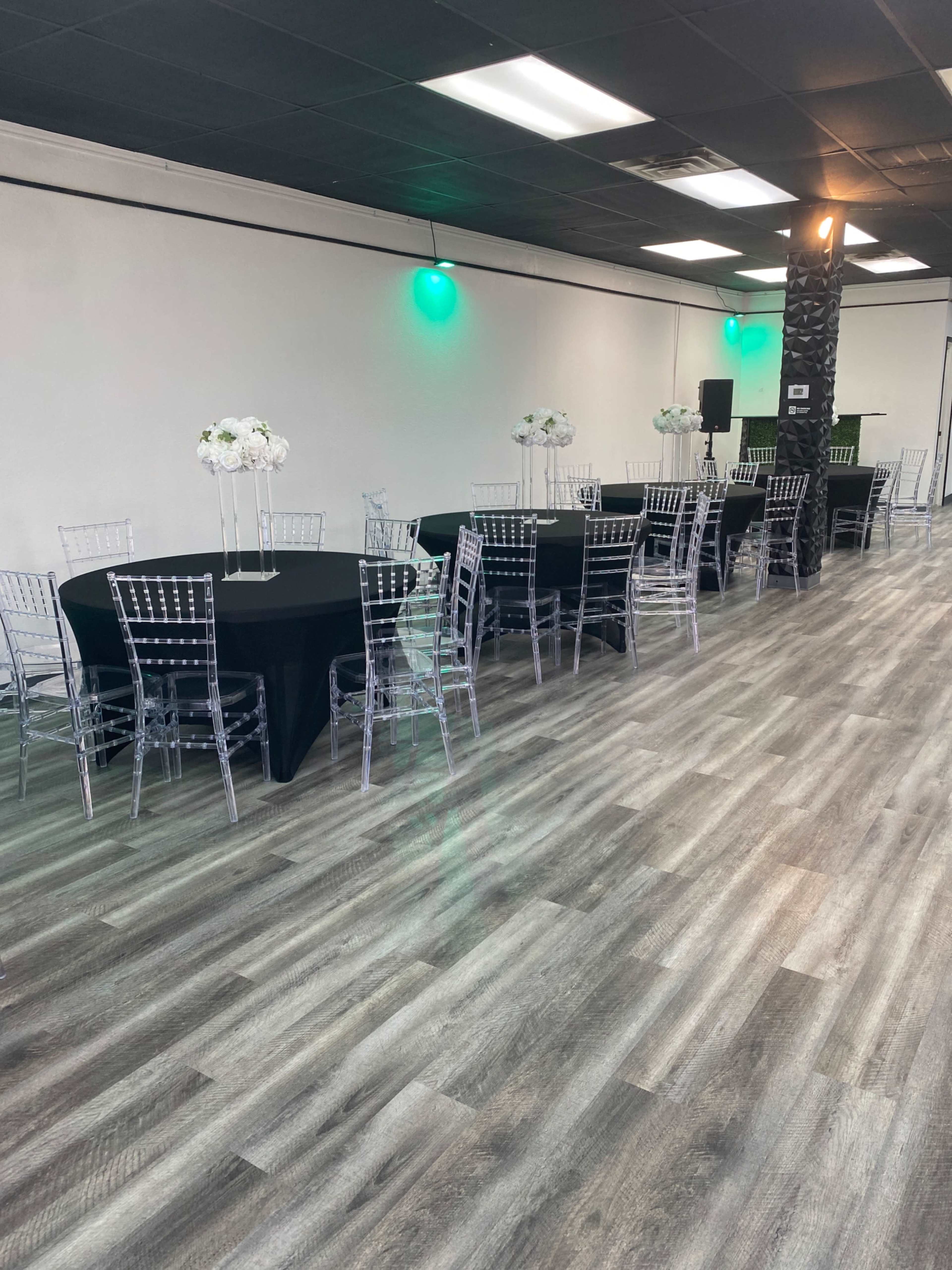 The image shows a banquet hall set up with round tables covered in black cloth and surrounded by clear plastic chairs, with floral centerpieces on each table.