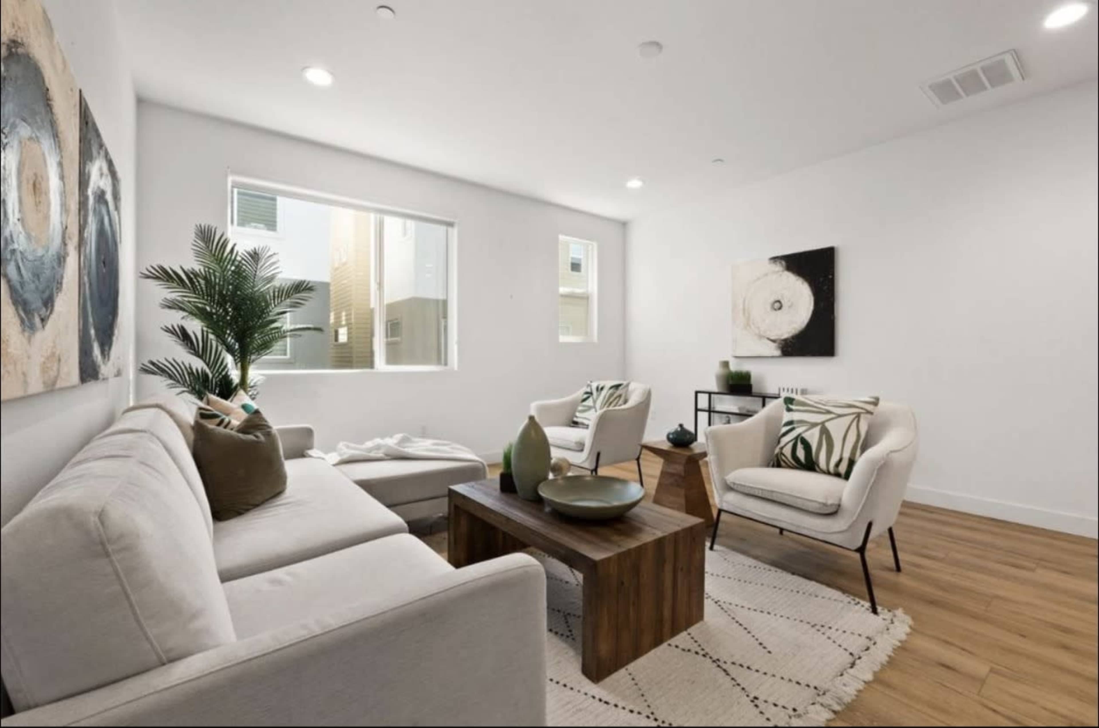 A bright living room features a light sofa and two armchairs arranged around a wooden coffee table, with simple artwork on the walls and a potted plant in the corner.