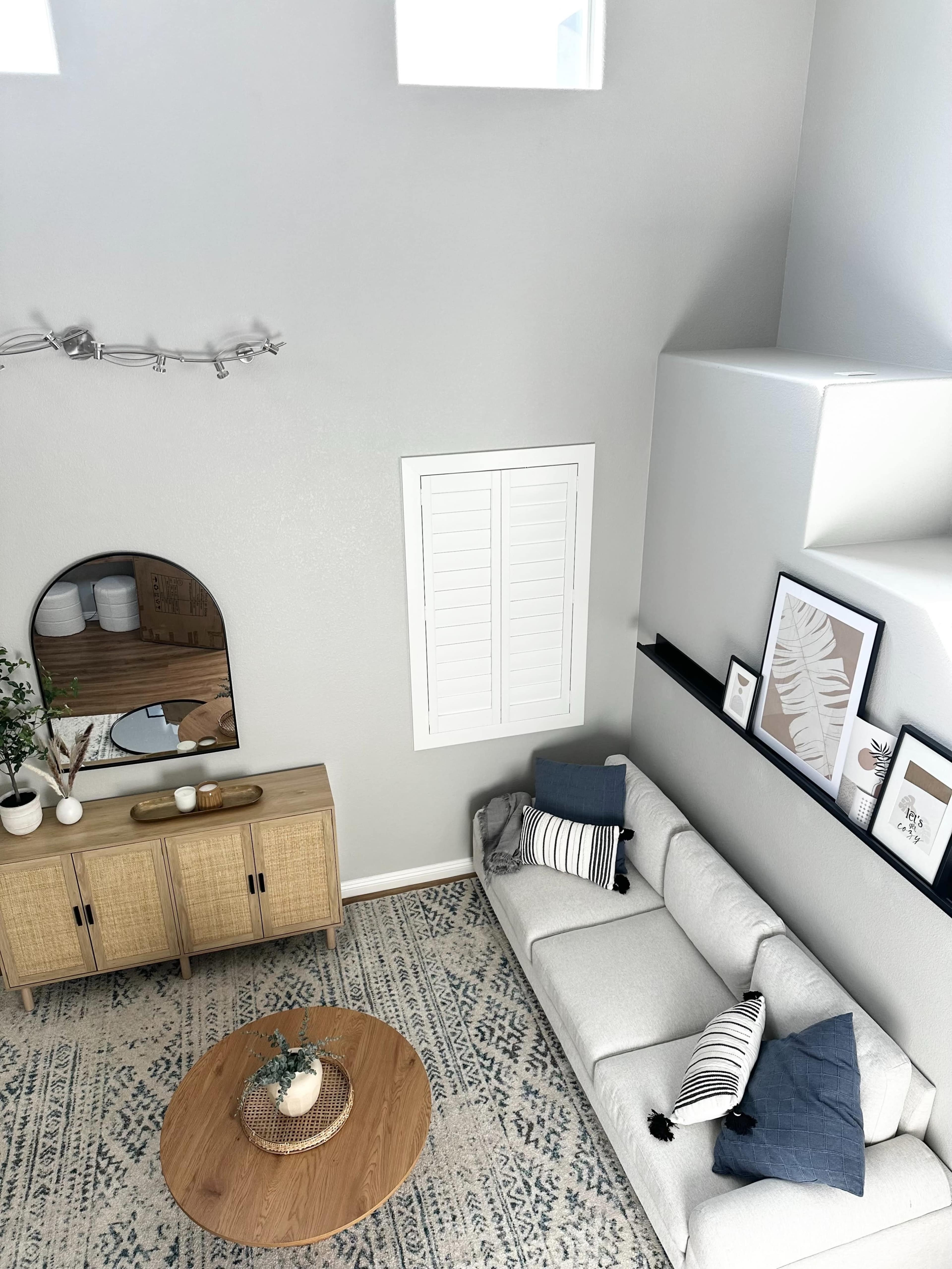 The image shows a modern living room featuring a light-colored sofa, a round wooden coffee table, a natural fiber area rug, and a decorative wall with framed art.