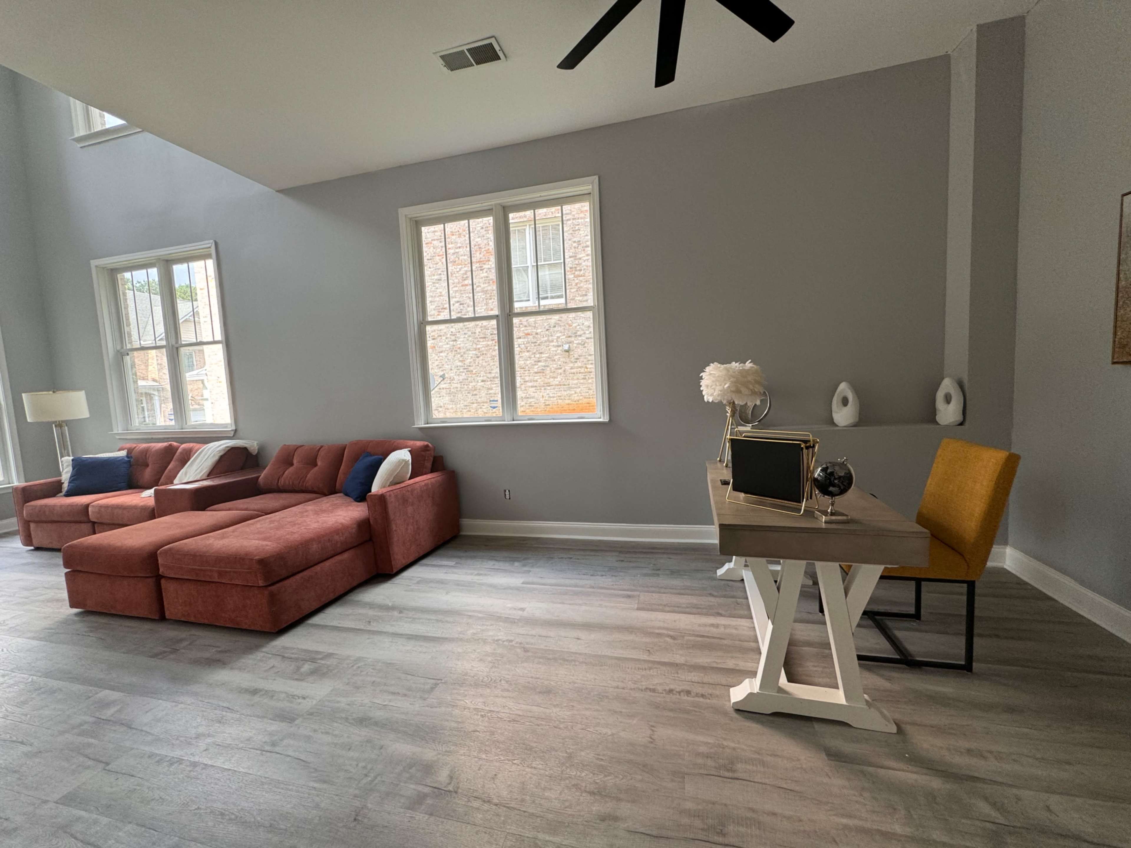 The image shows a spacious living area featuring a red sectional sofa, a wooden desk with a yellow chair, and large windows allowing natural light to enter.