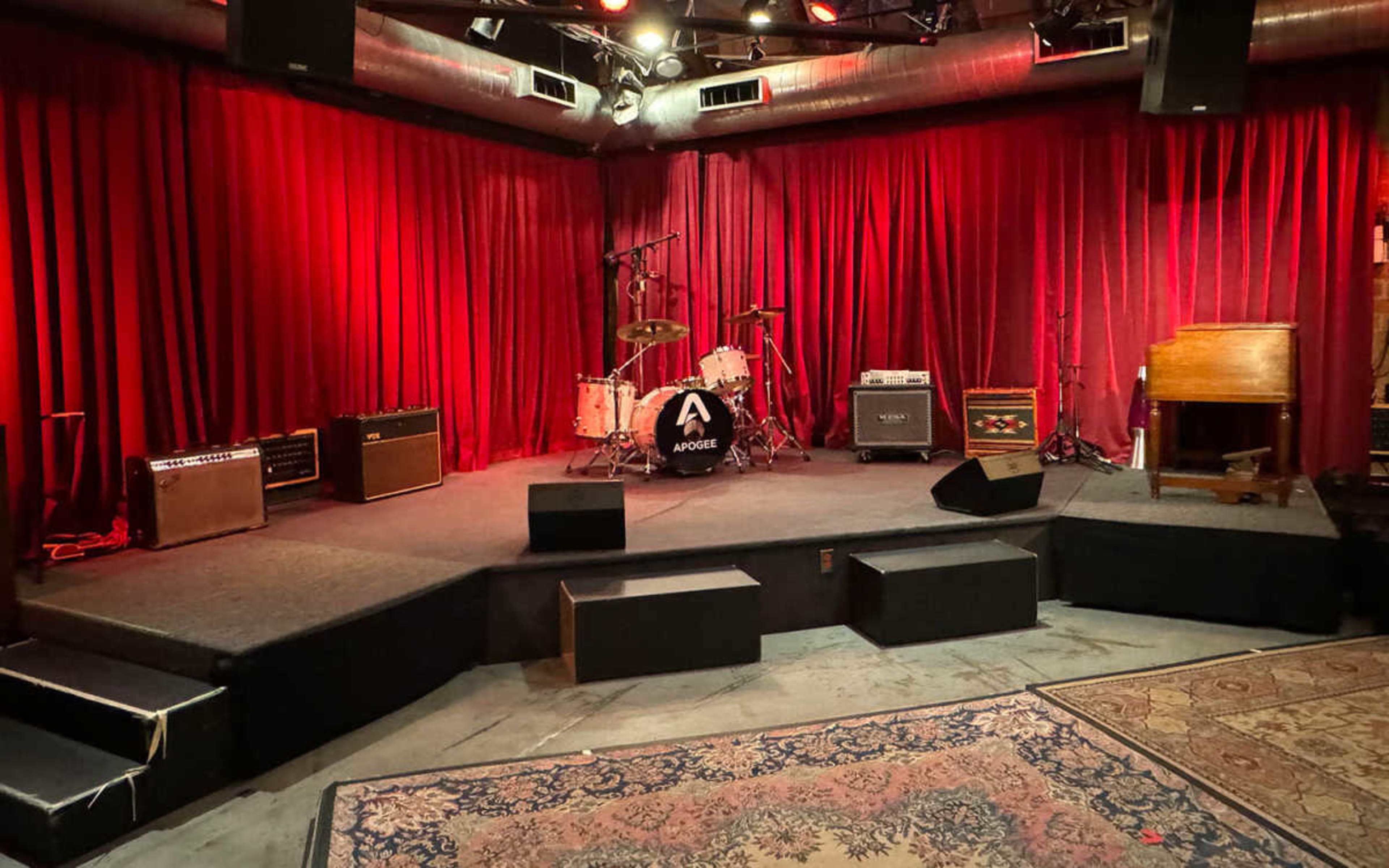 The image shows a music stage setup, featuring a drum kit in front of red curtains, amplifiers on the sides, and a vintage keyboard on one side.