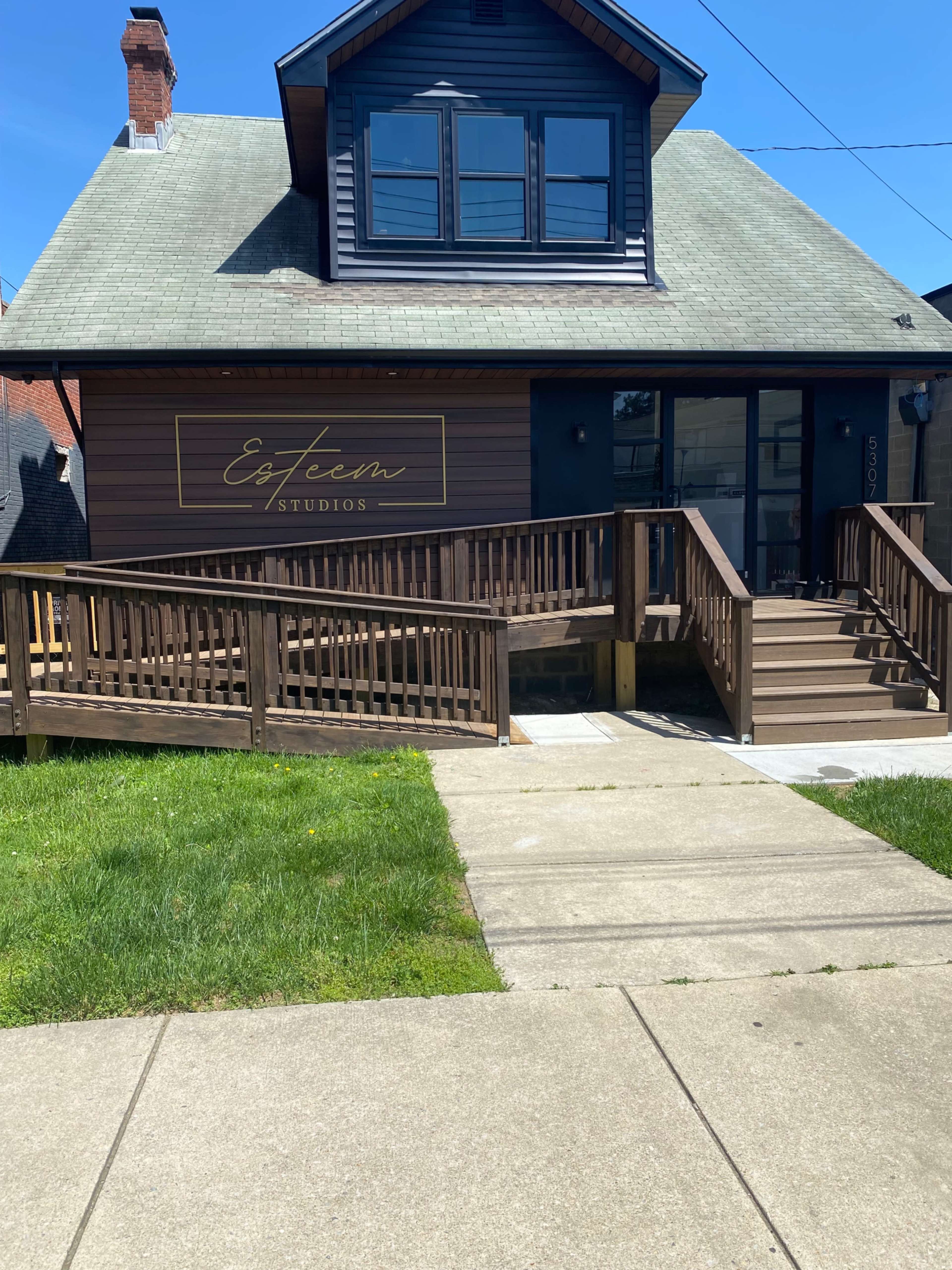 The image shows a wooden house with a sloped roof and a sign for "Esteem Studios" on the front, featuring a ramp and stairs leading to the entrance.