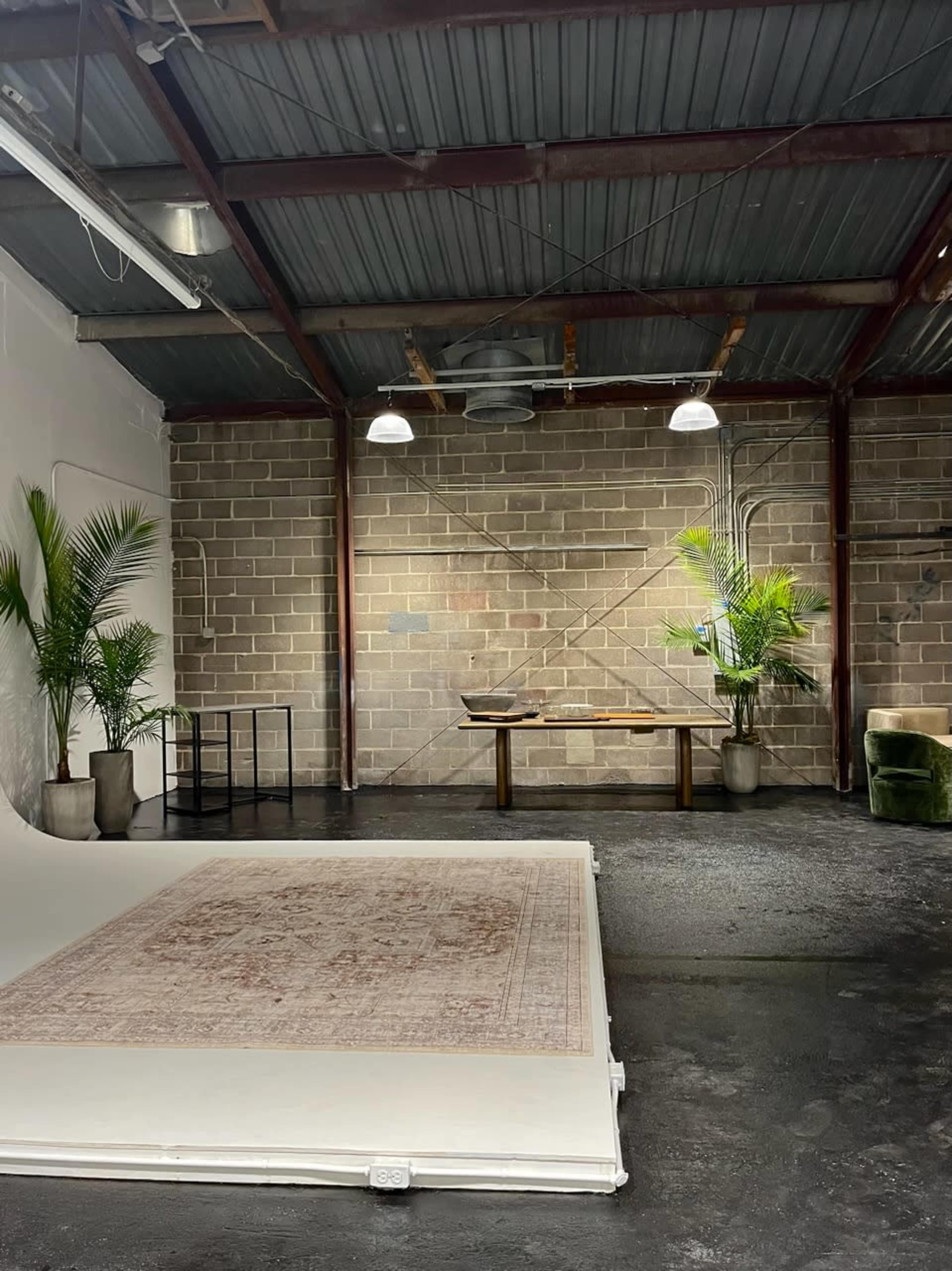 A spacious industrial-style room with a rug, a wooden table, potted plants, and exposed brick walls.