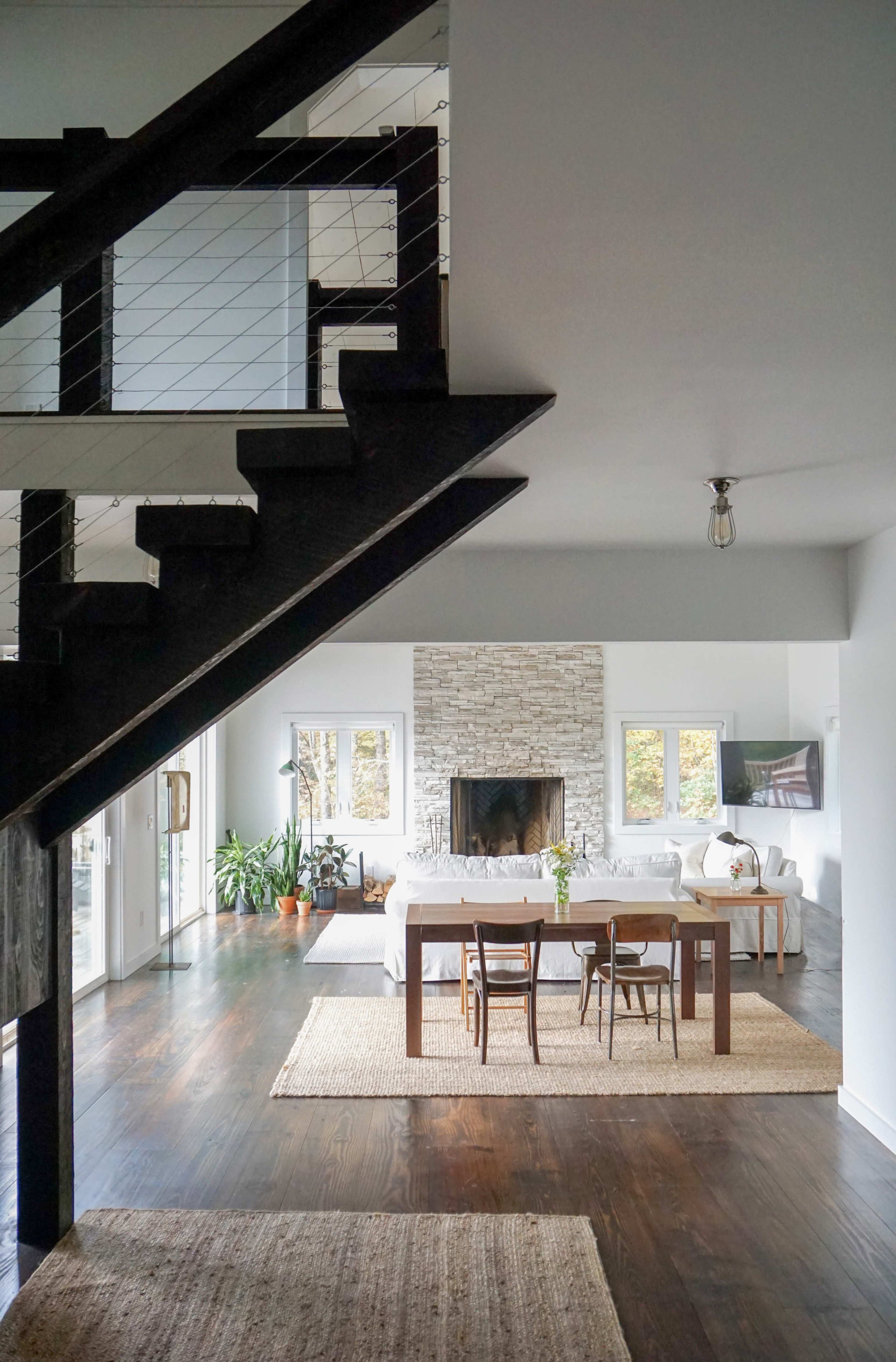 A modern living room features a stone fireplace, dining area with a wooden table, and large windows allowing natural light.