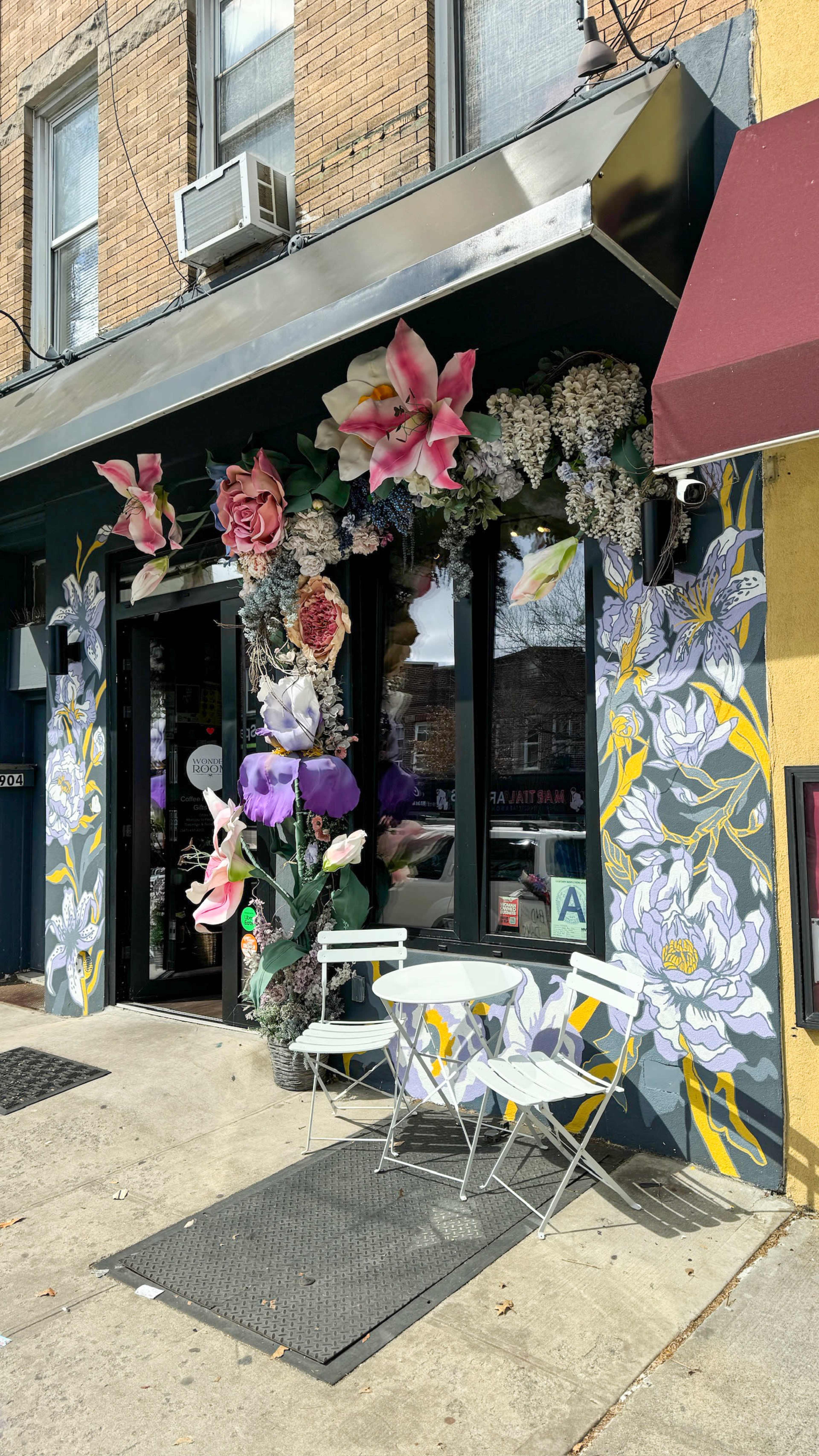 A storefront is adorned with large floral murals and decorations, featuring a small white table and chairs outside.