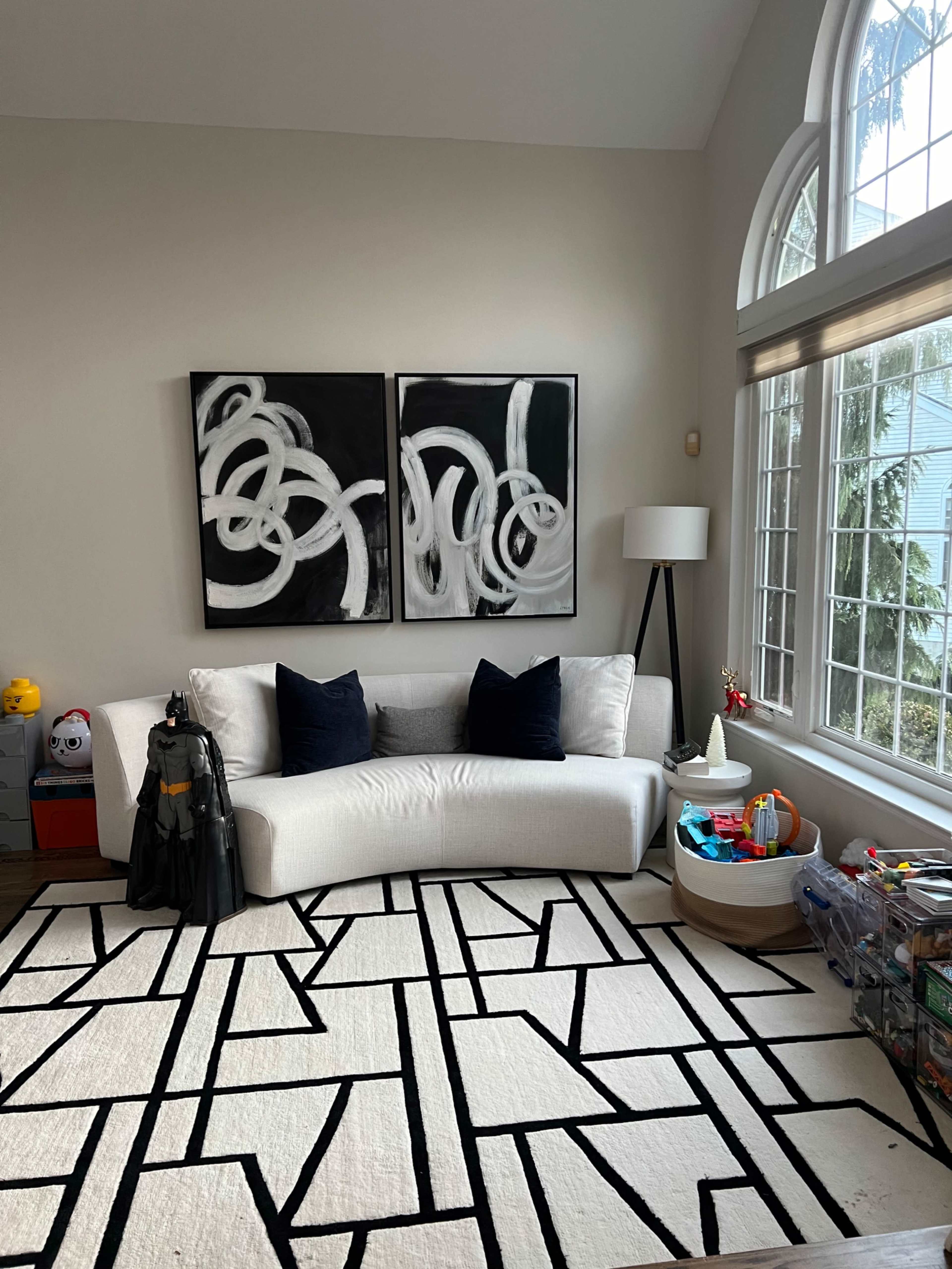 The image shows a modern living room with a white curved sofa, two abstract black-and-white paintings on the wall, and a large rug with a geometric pattern.