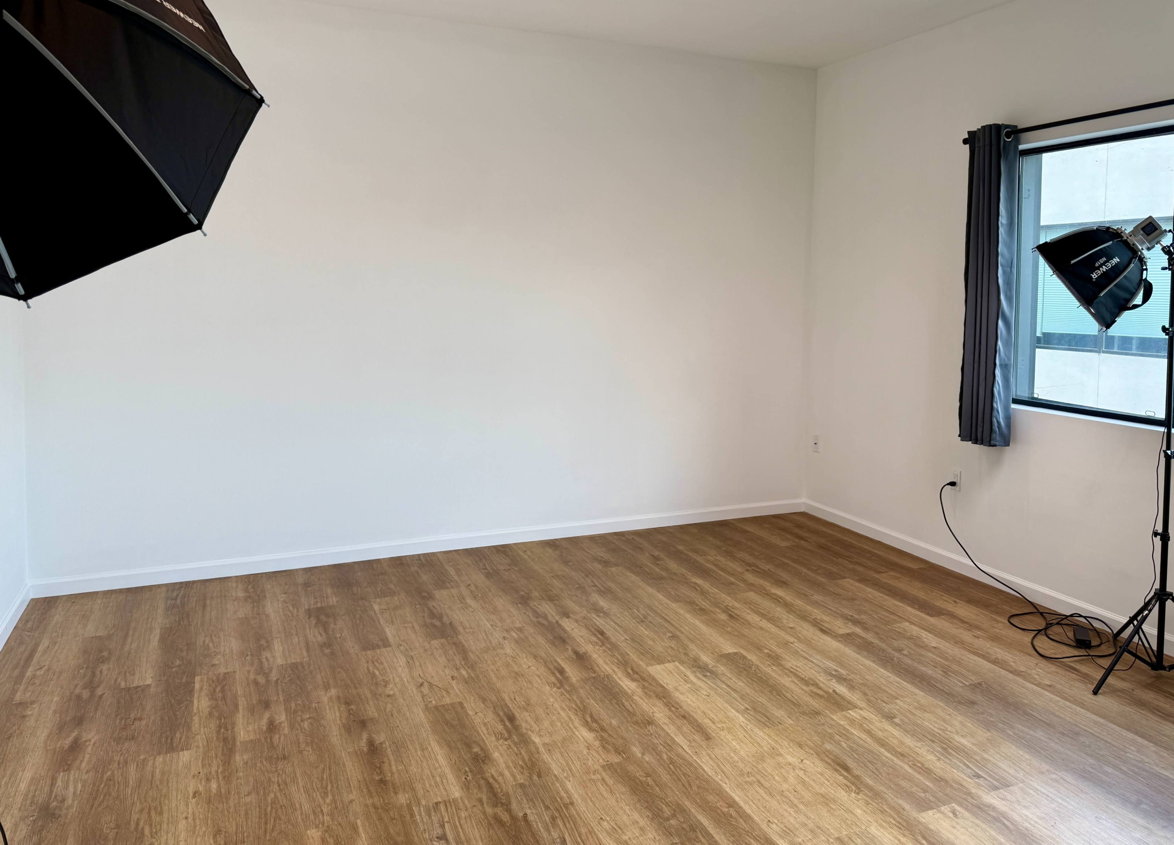 The image shows a vacant room with wooden flooring, a white wall, and two lighting setups in the corners.