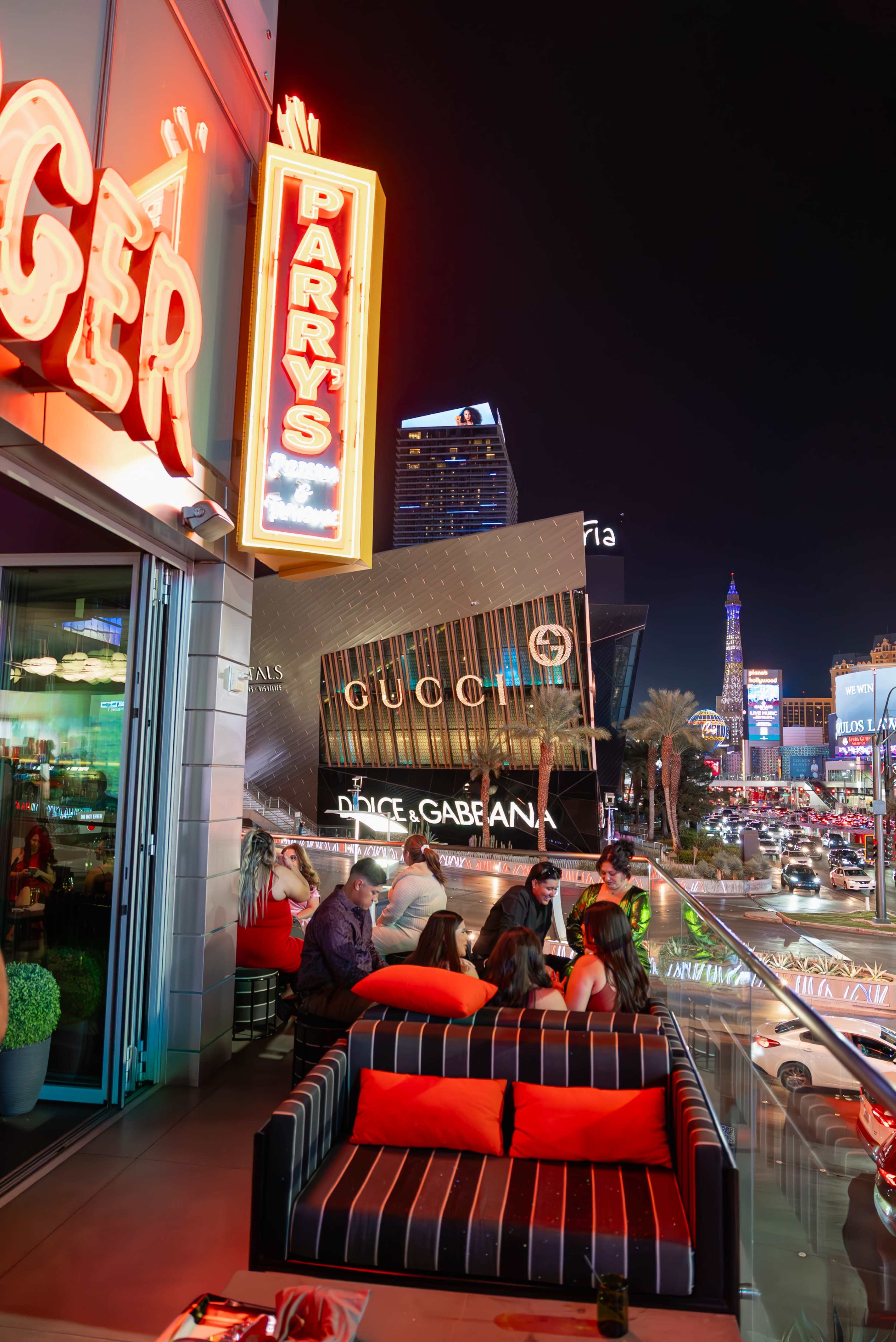 The image shows a brightly lit outdoor dining area in Las Vegas, featuring a neon sign for "Parry's" and visible views of high-rise buildings and the bustling nightlife.