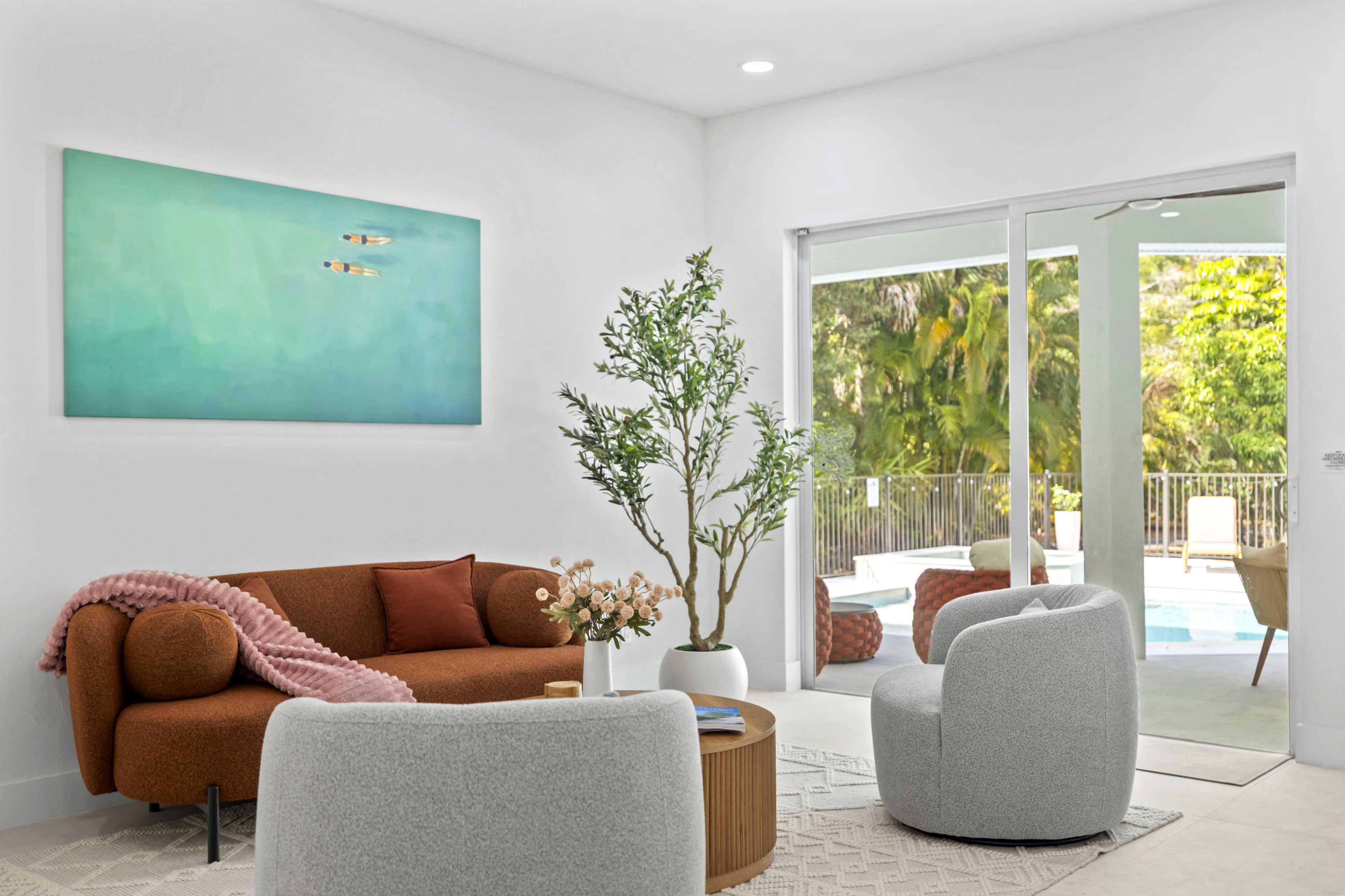 A modern living room features a brown sofa, two light gray chairs, a round coffee table, a large potted plant, and a view of a pool area through sliding glass doors.
