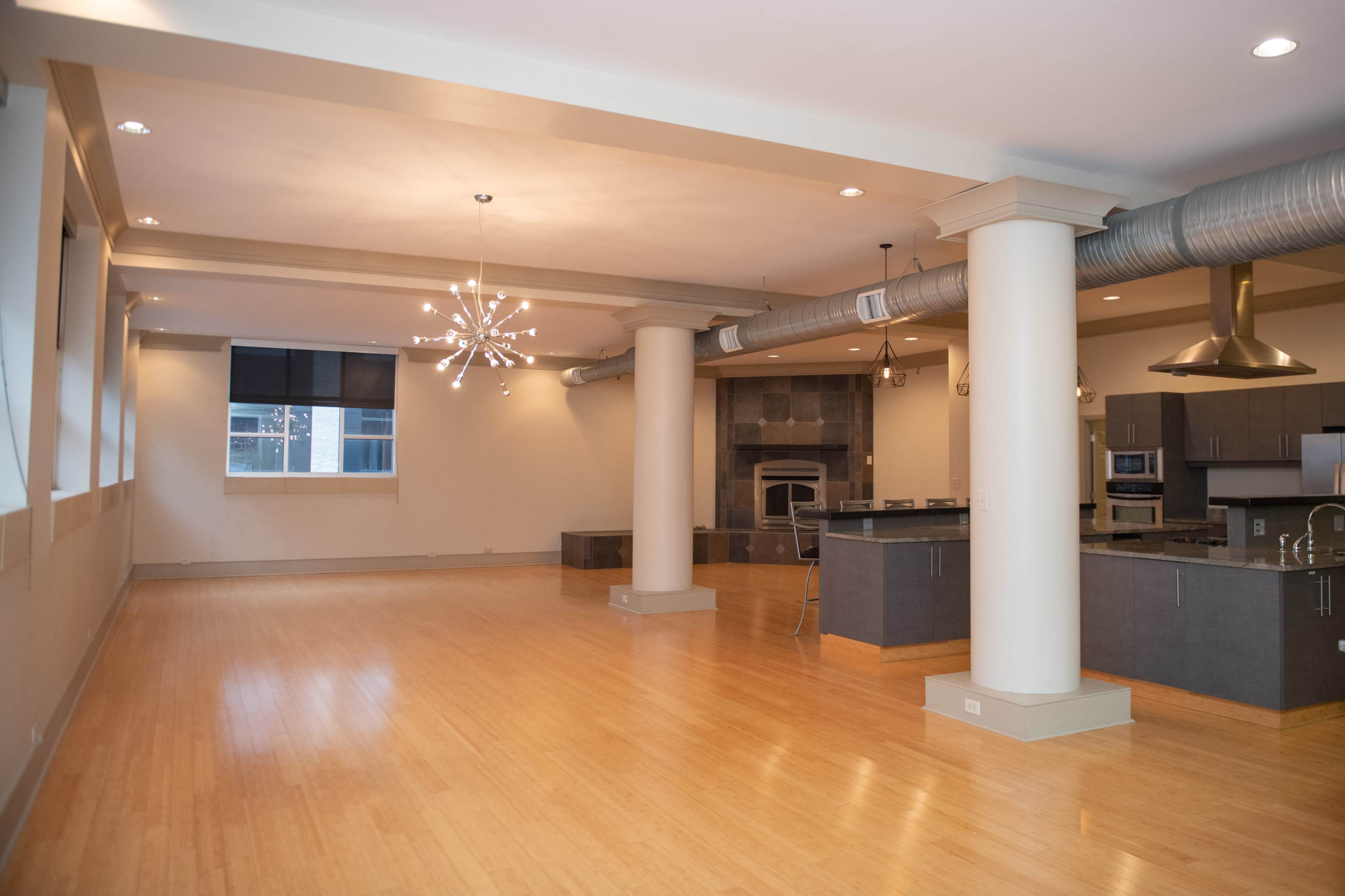 The image shows a spacious, modern open-concept living area with wooden flooring, large windows, and a kitchen featuring dark cabinetry and stainless steel appliances.