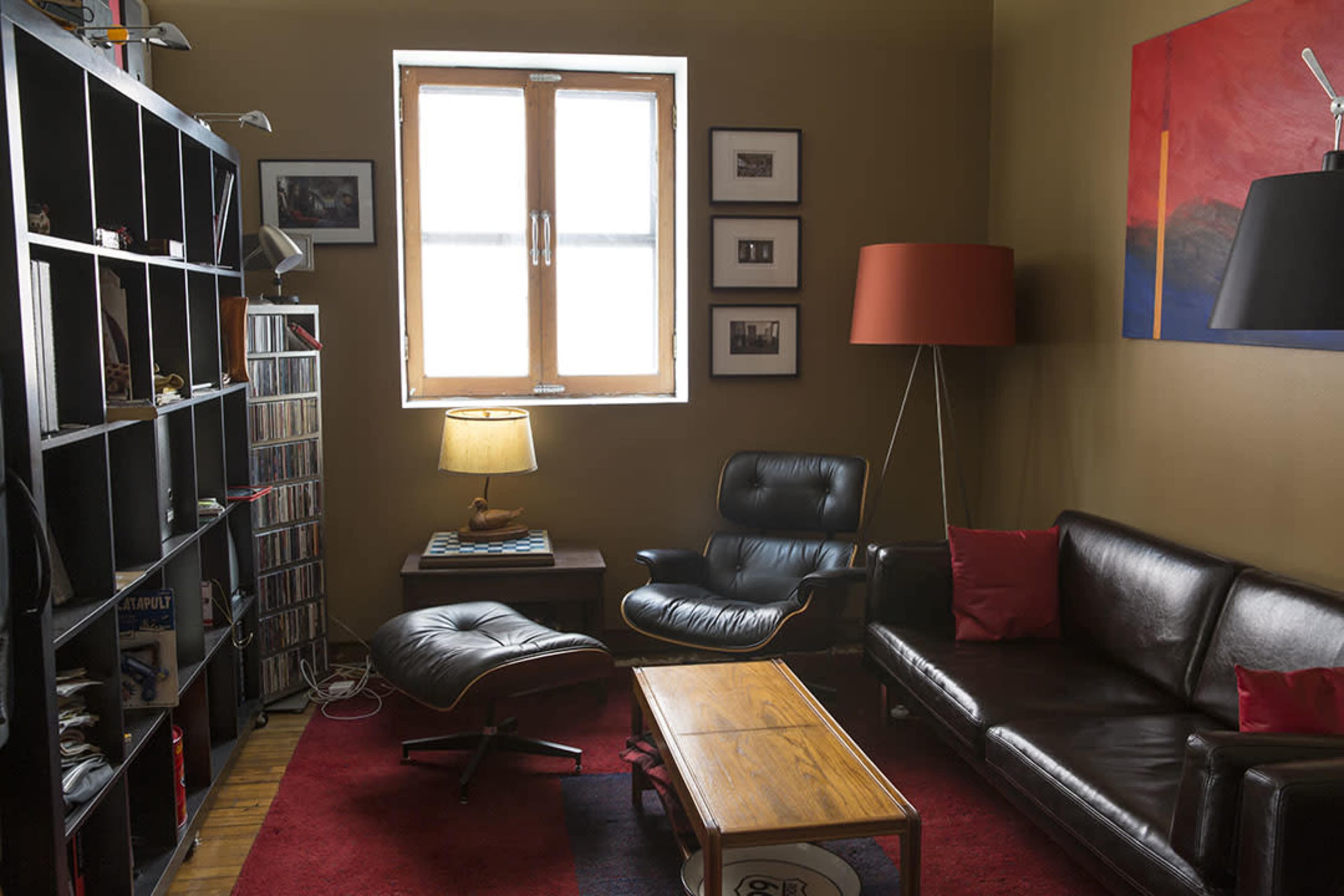A cozy living room with a black leather sofa, a wooden coffee table, a large bookshelf filled with CDs, and a window illuminating the space.
