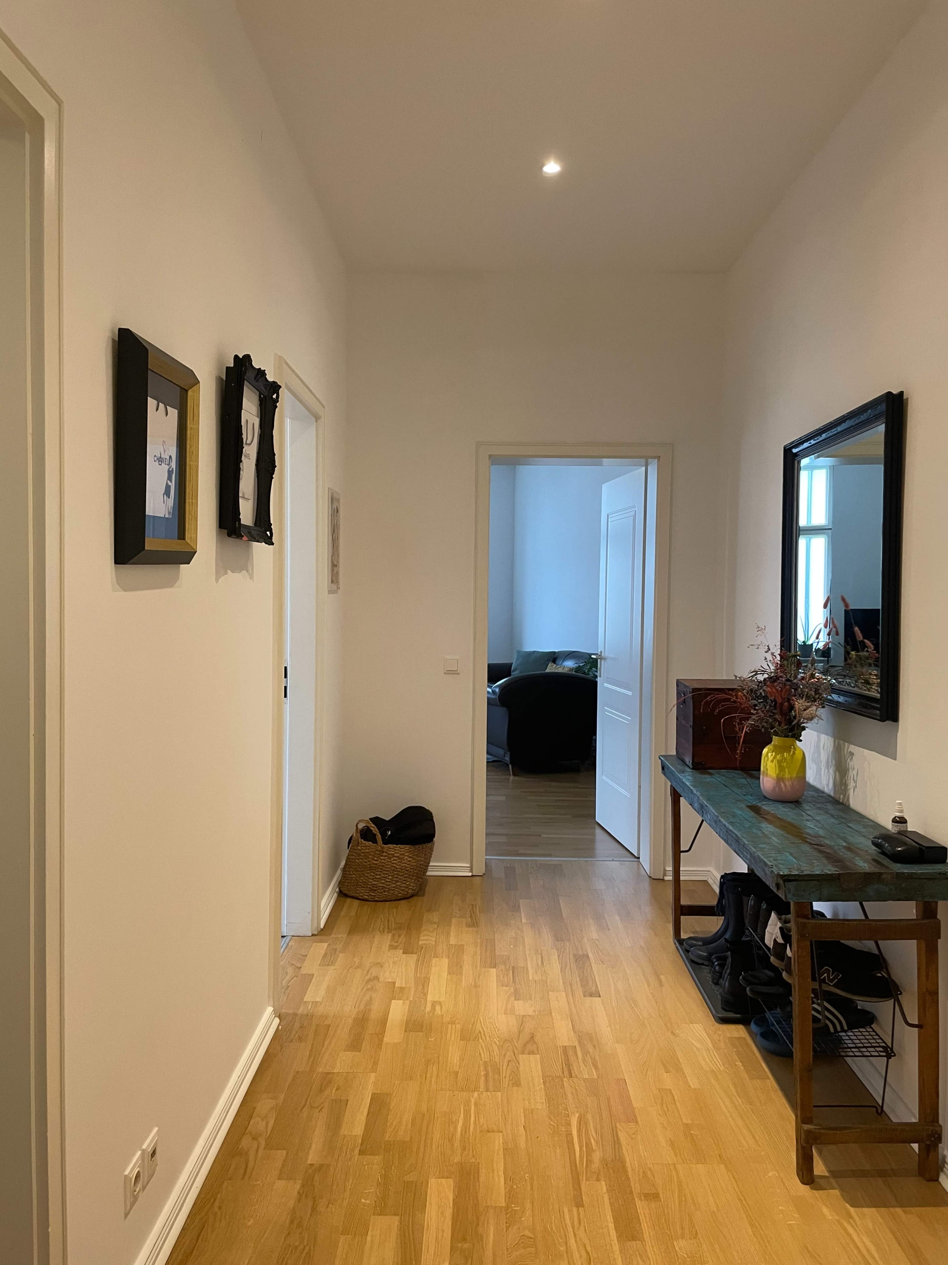 The image shows a hallway with wooden flooring, framed pictures on the walls, a console table, and an open doorway leading to a room in the background.