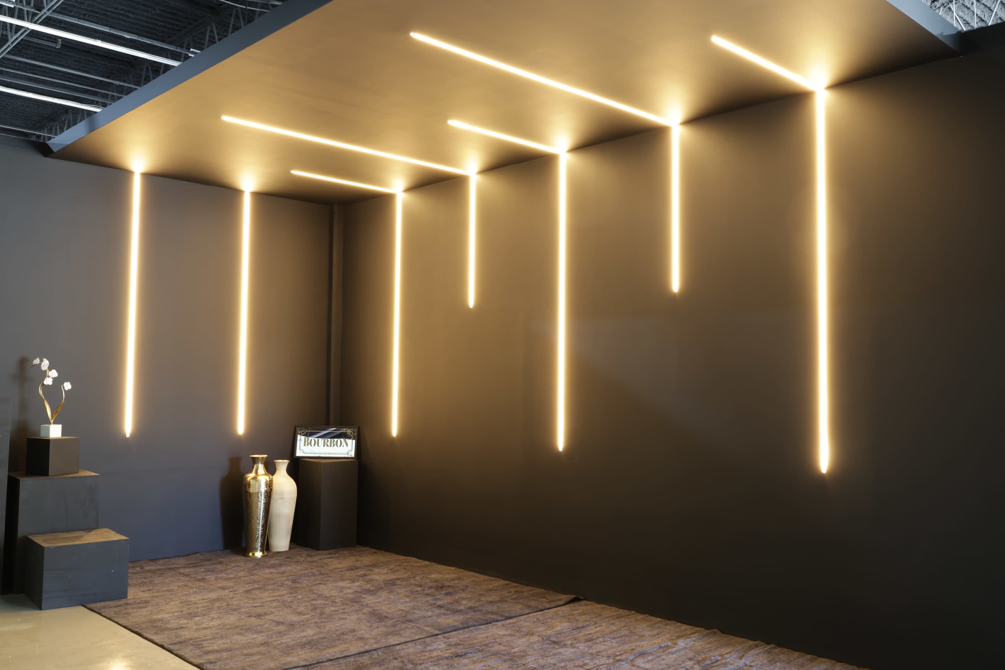 The image shows a modern interior space with vertical light fixtures illuminating a black wall, complemented by a decorative vase on a pedestal and a rug on the floor.