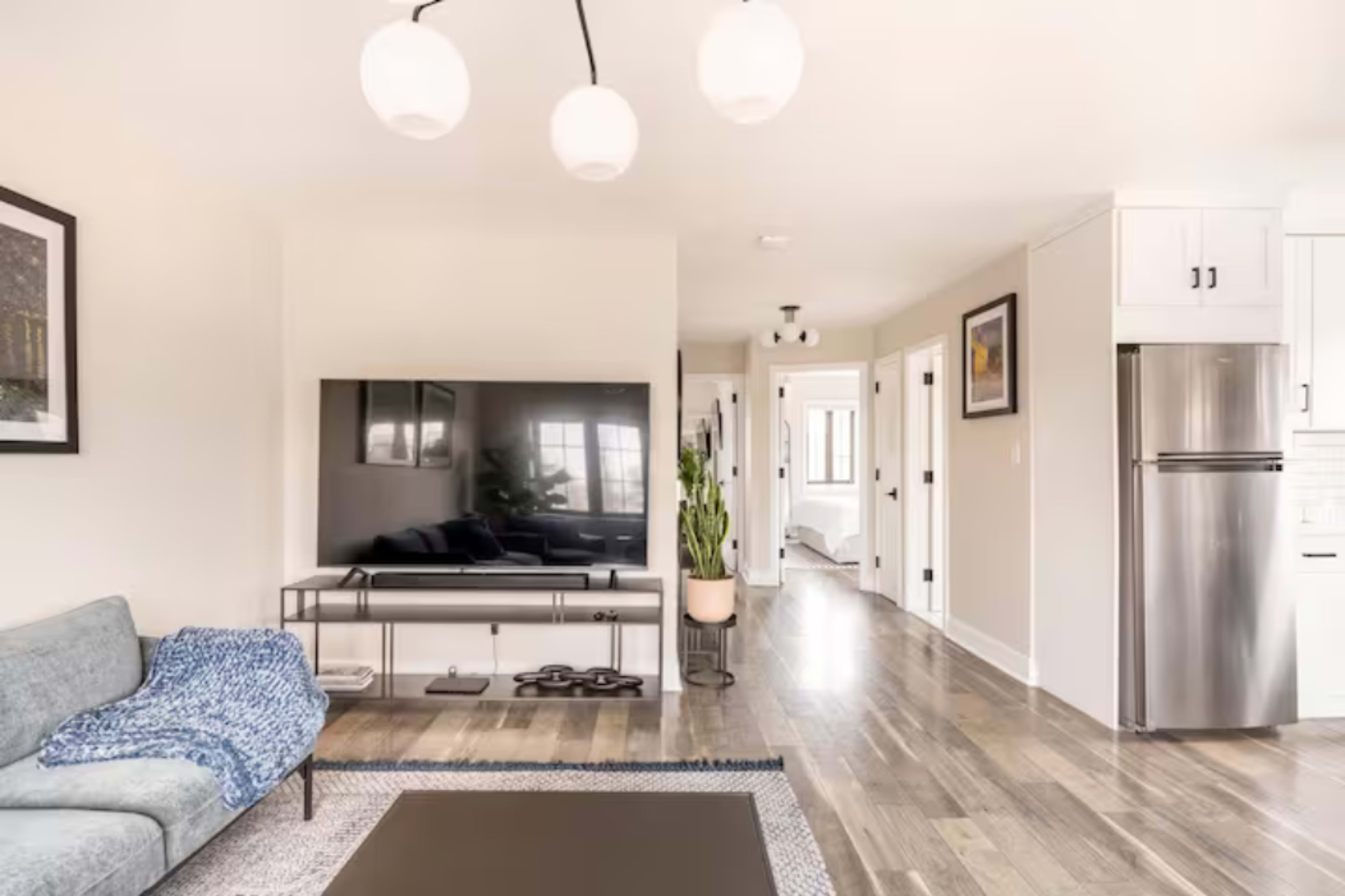 A modern living room features a gray sofa, a large flat-screen TV on a stand, and a view of a hallway leading to additional rooms.