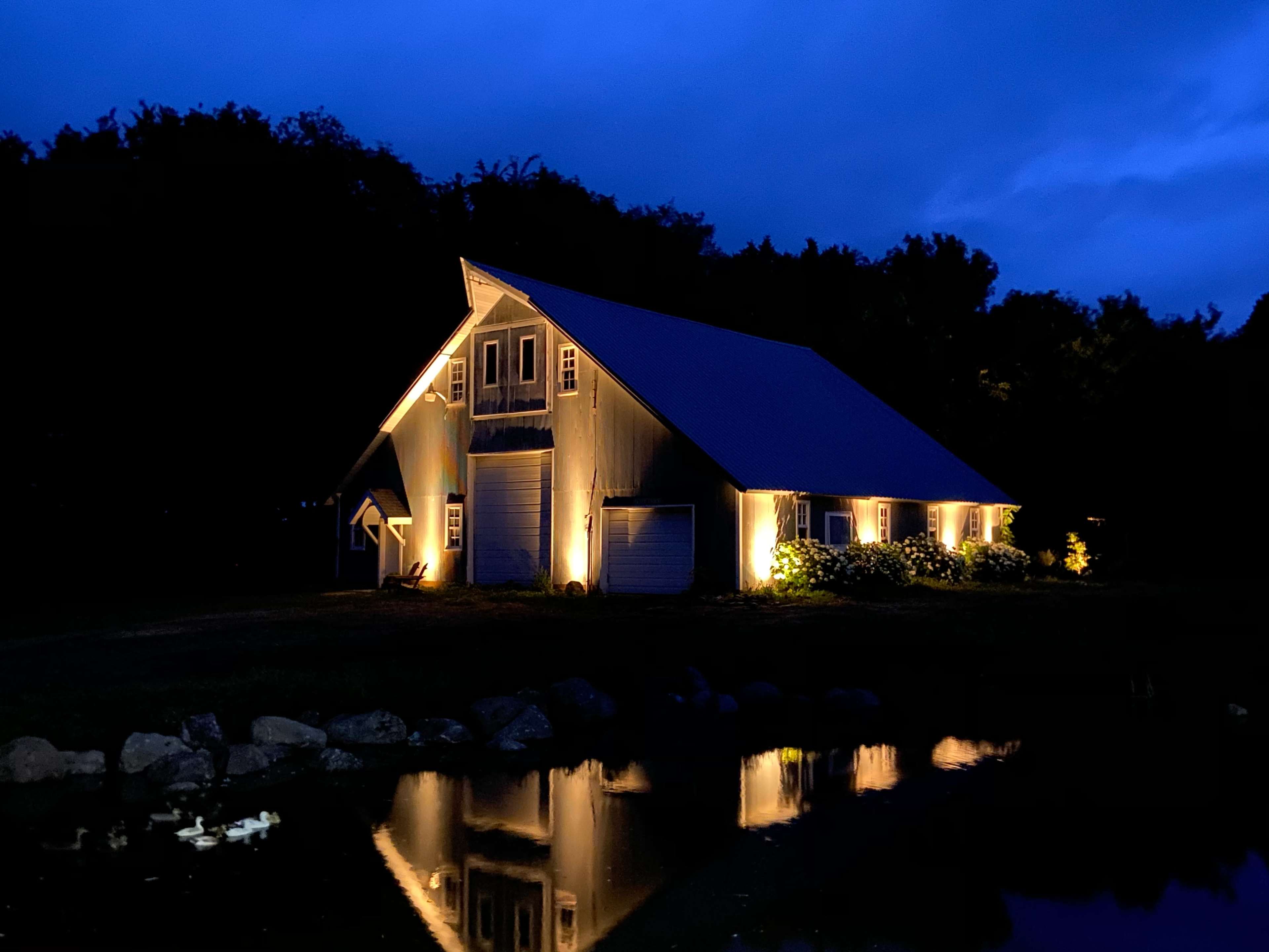 A barn-like structure is illuminated by warm lights, reflecting in a calm body of water under a twilight sky.