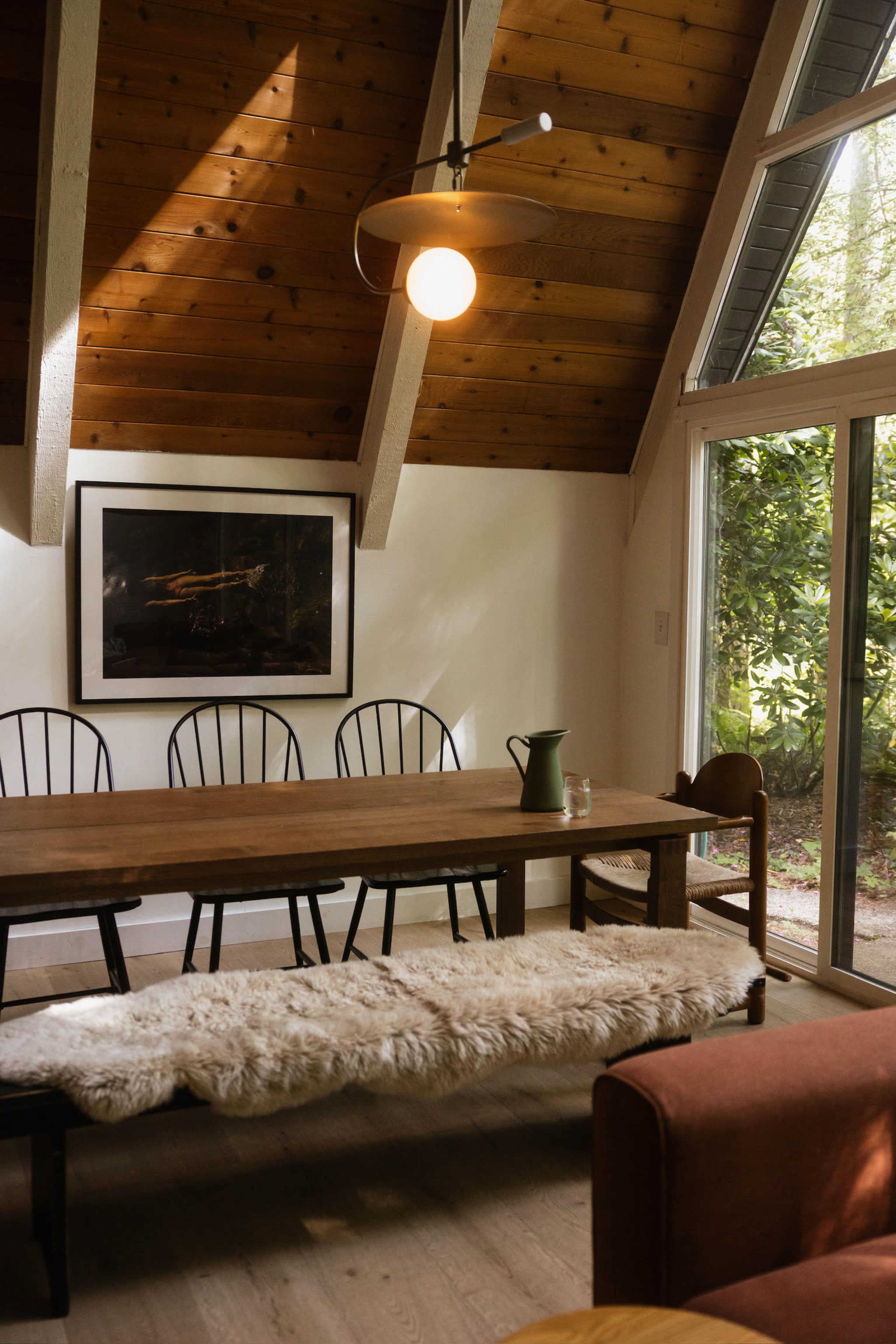 The image depicts a cozy dining area featuring a wooden table with black chairs, a bench with a fur throw, and large windows that allow natural light to illuminate the space.