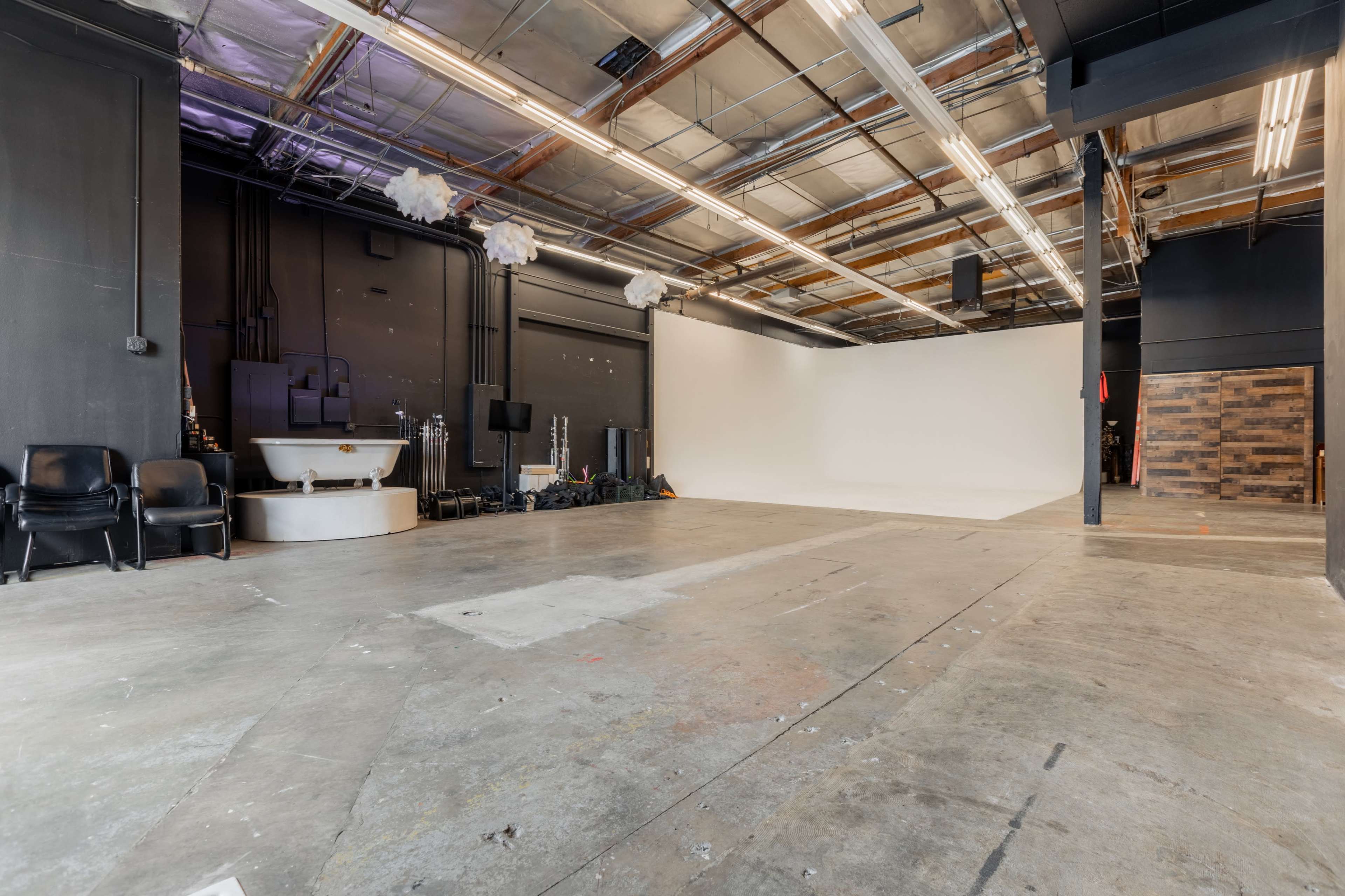 The image shows a spacious photography studio featuring a plain white backdrop, a concrete floor, and several studio lights and equipment in the background.