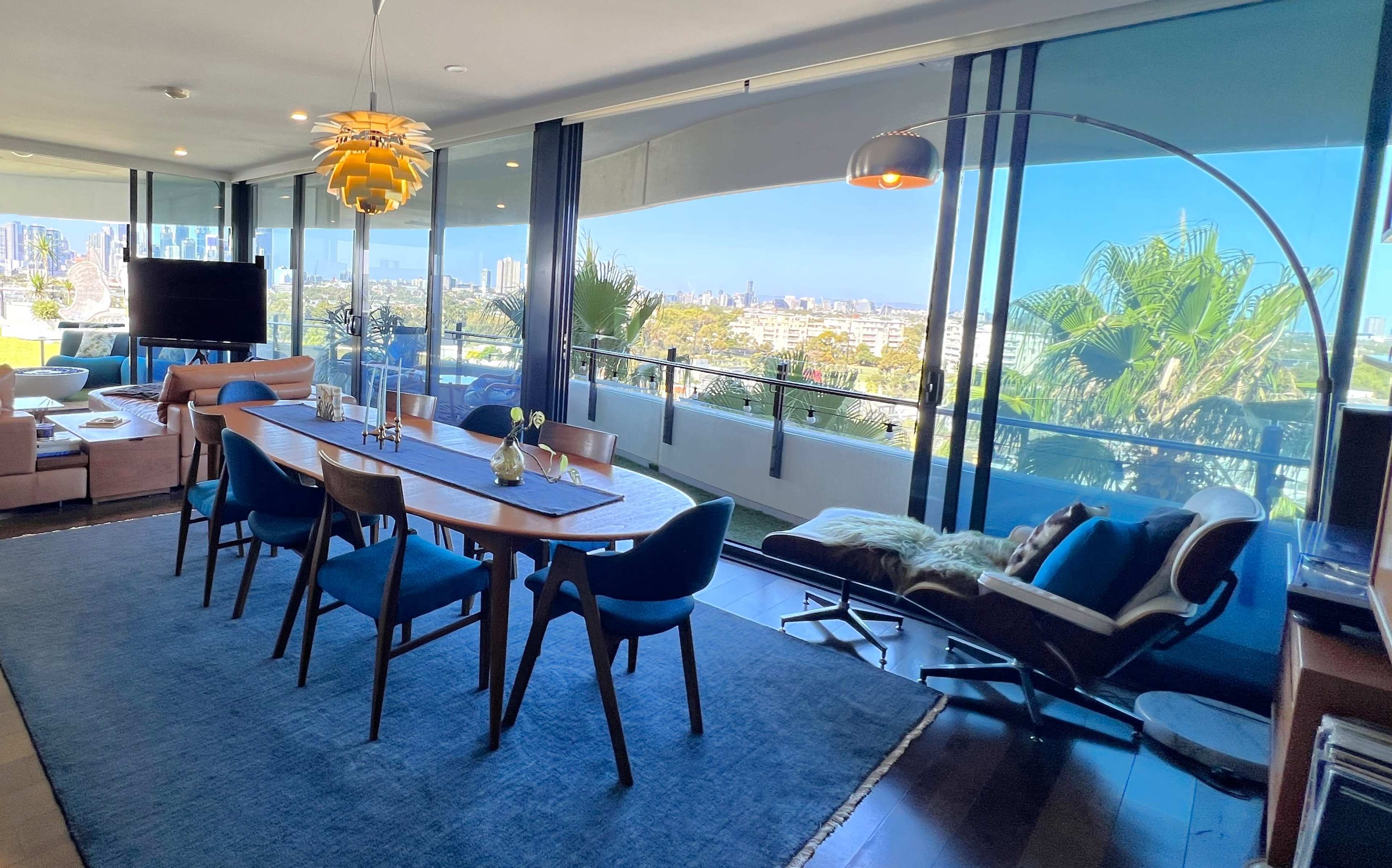A modern dining area features a long table with blue chairs, an arc floor lamp, and a view of a city skyline through large glass windows.