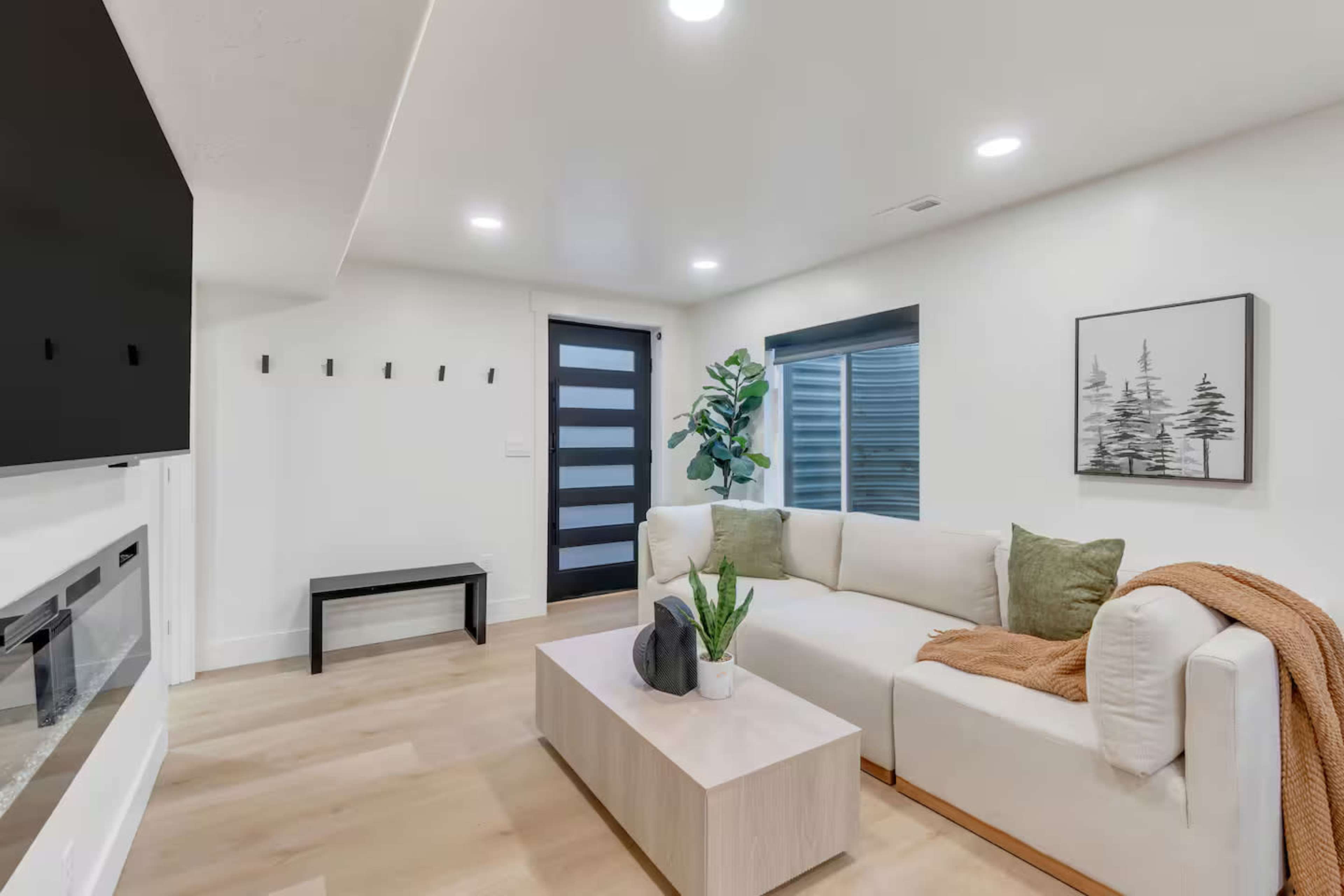 A modern living room features a light-colored sectional sofa, a wooden coffee table, and a potted plant, with a black accent wall and a front door with horizontal glass panels.