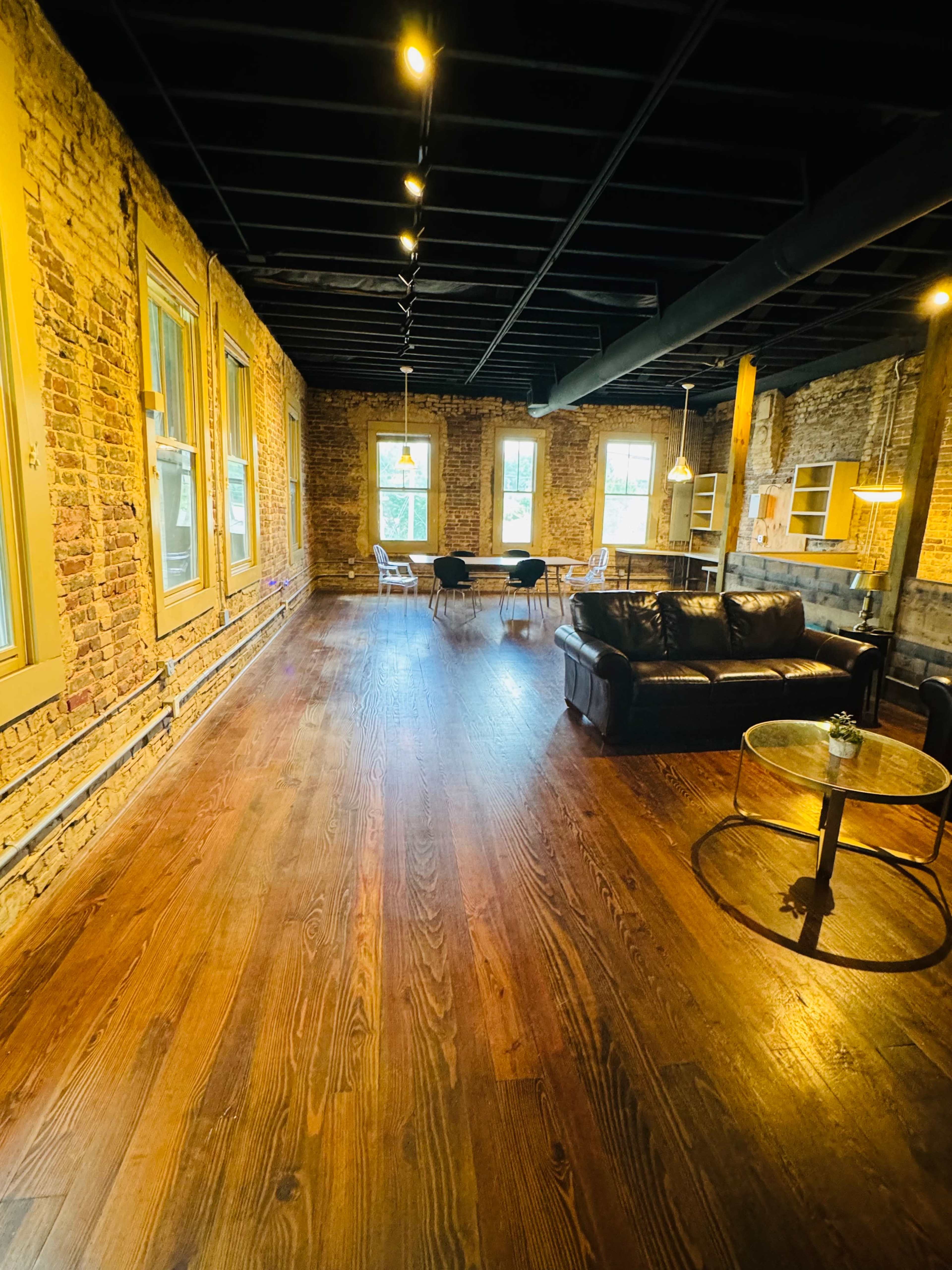 An open, rustic-style room featuring exposed brick walls, wooden floors, and a modern seating area with a black leather couch and a circular coffee table.