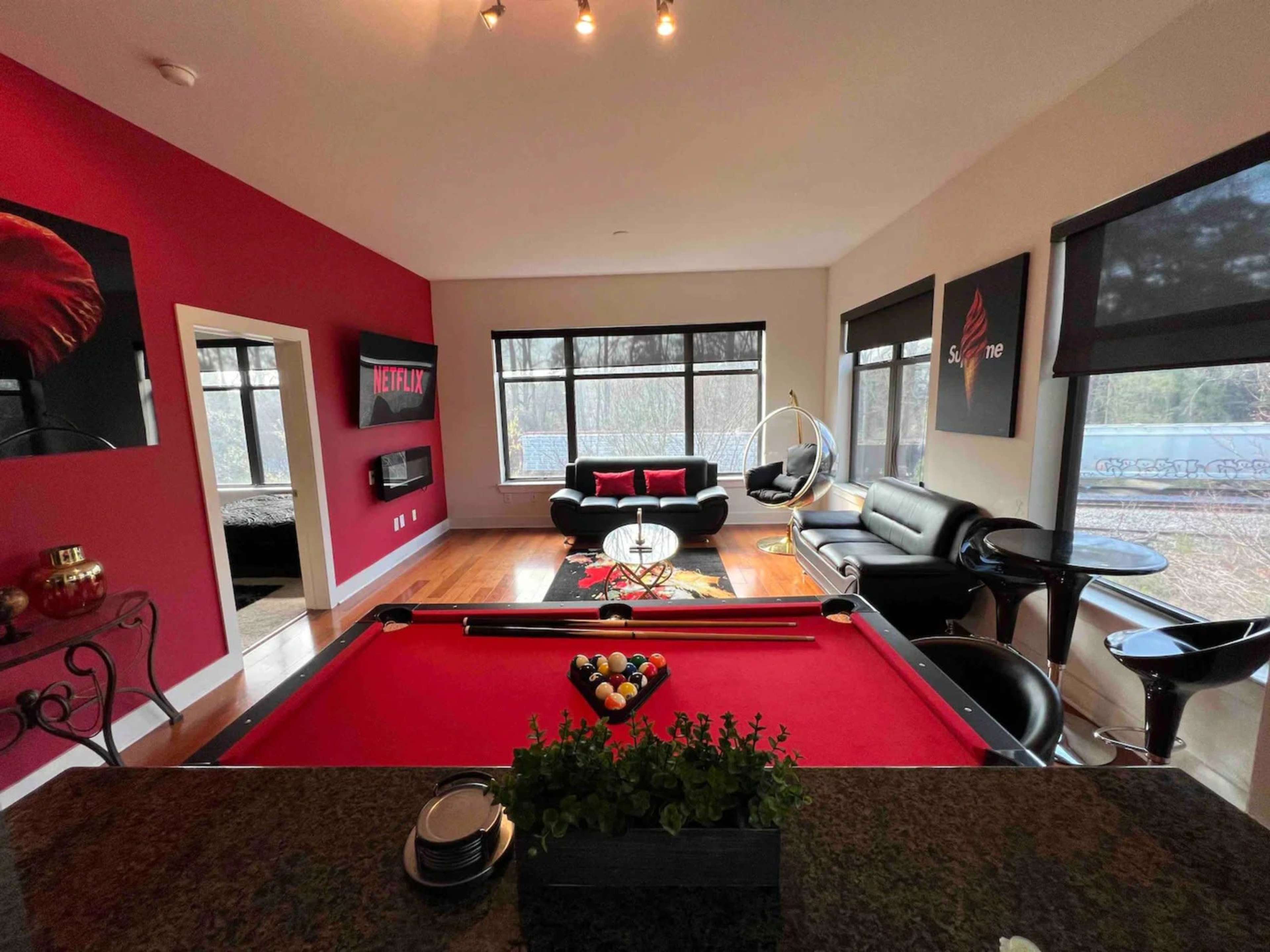 A modern living space featuring a pool table at the forefront, a seating area with black sofas, and large windows providing natural light.