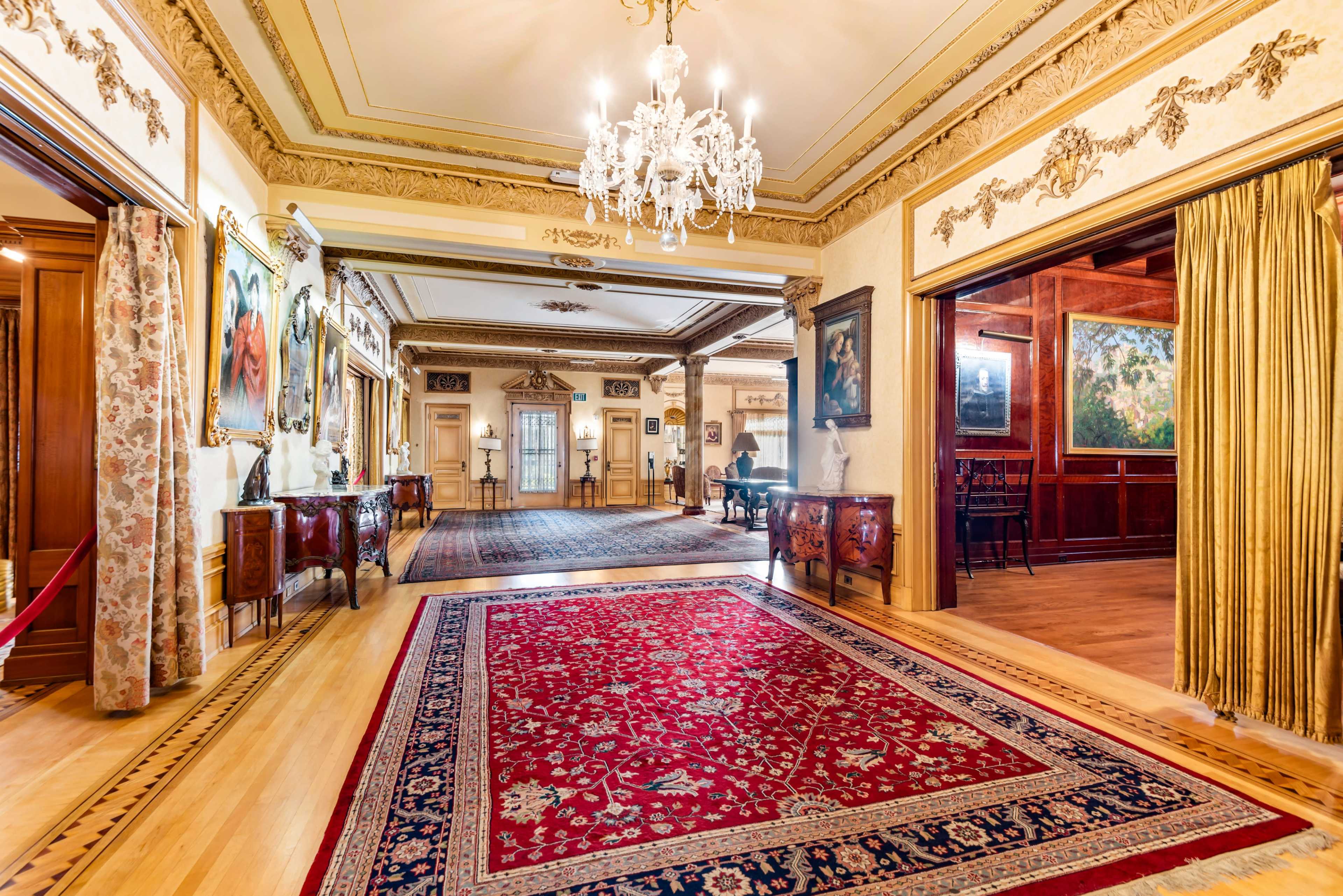 The image shows an ornate hallway in a historic building featuring intricate moldings, framed paintings on the walls, a chandelier, and a large area rug.