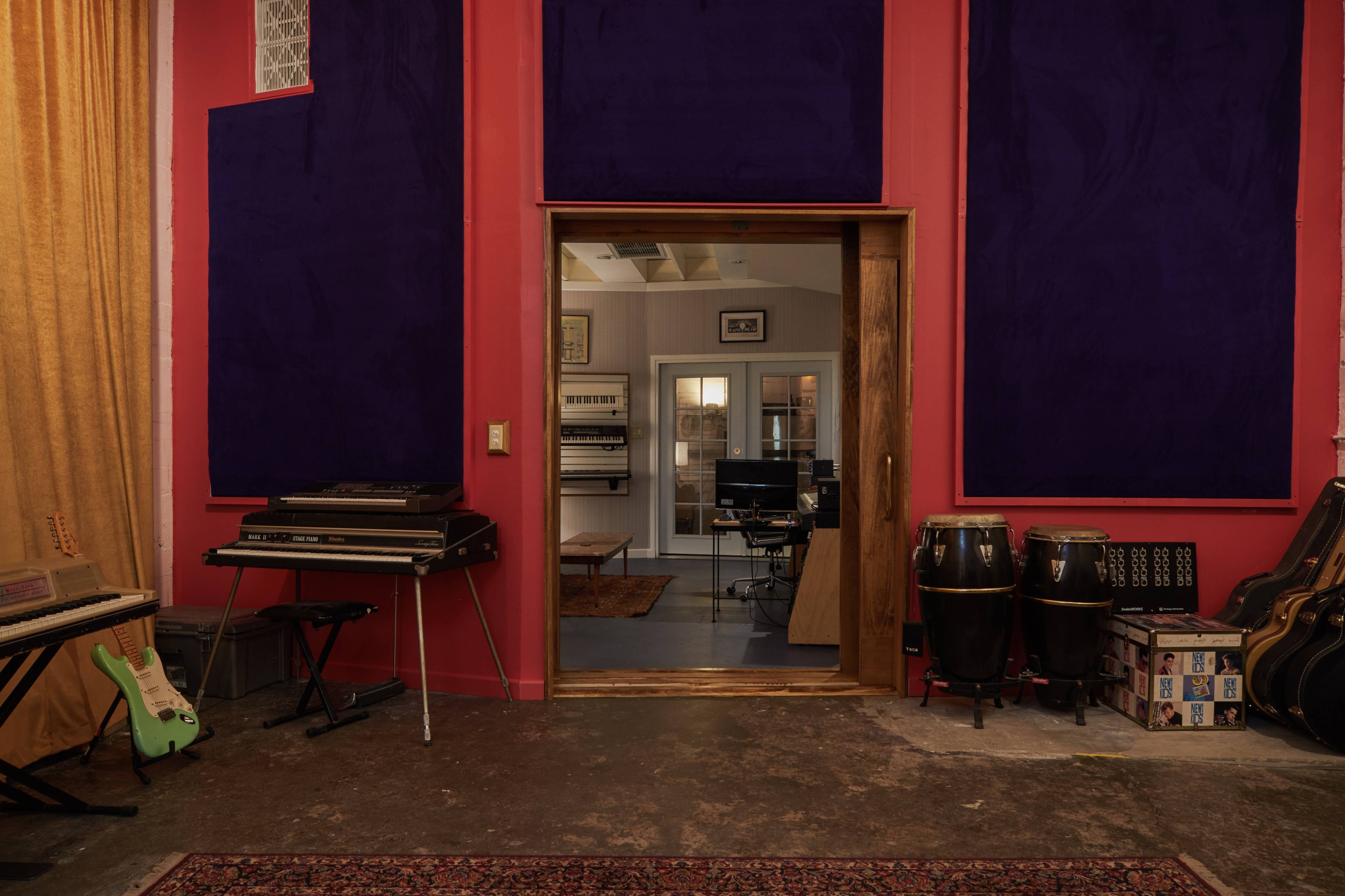 The image shows a music studio with colorful walls, featuring a doorway leading to a space equipped with instruments and recording equipment.