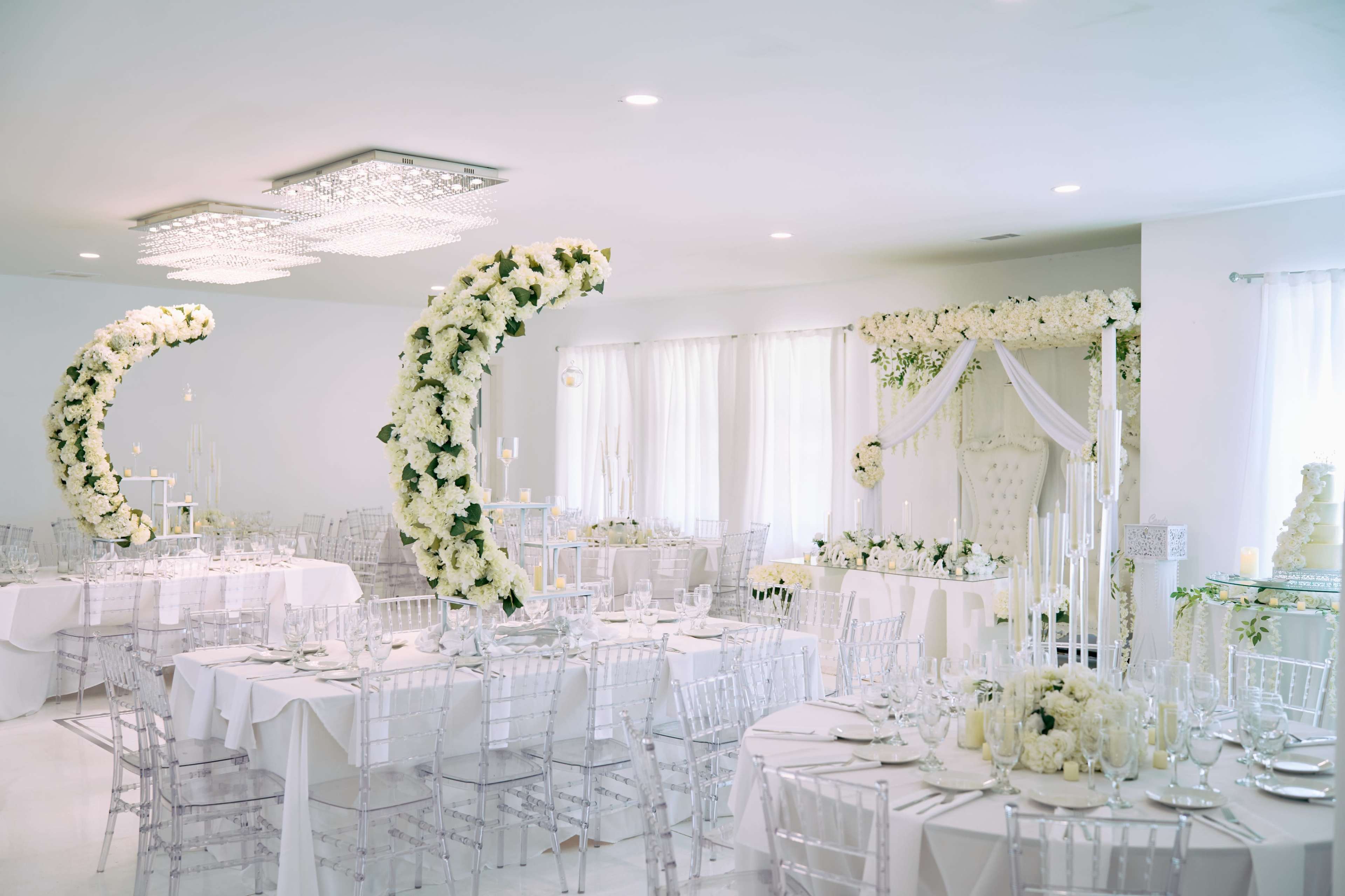 The image shows an elegantly decorated banquet hall with white tablecloths, clear chairs, and large floral arrangements shaped like crescent moons.