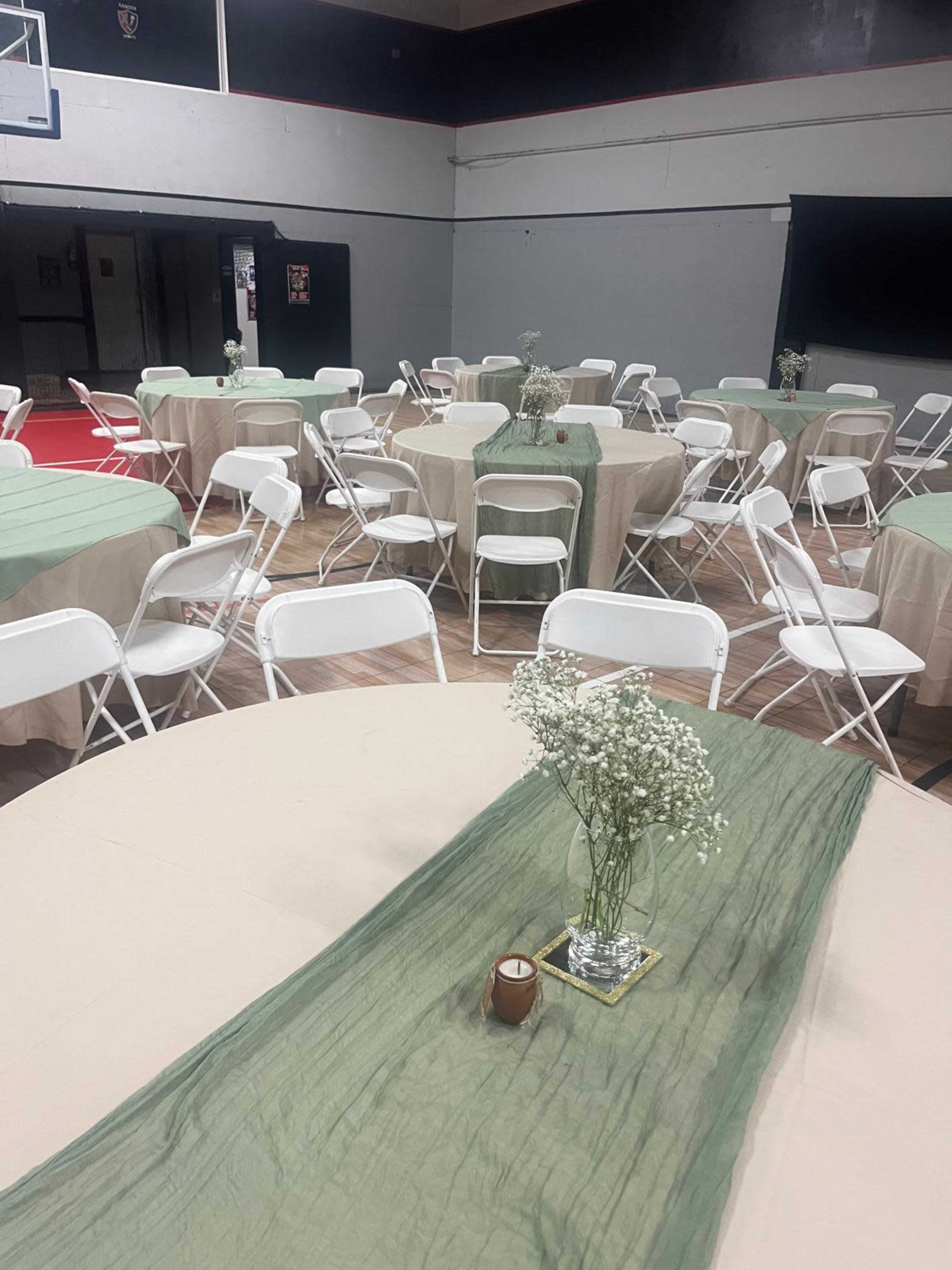 The image shows a gymnasium set up for an event, featuring round tables covered with beige tablecloths and green runners, surrounded by white folding chairs and adorned with floral centerpieces.