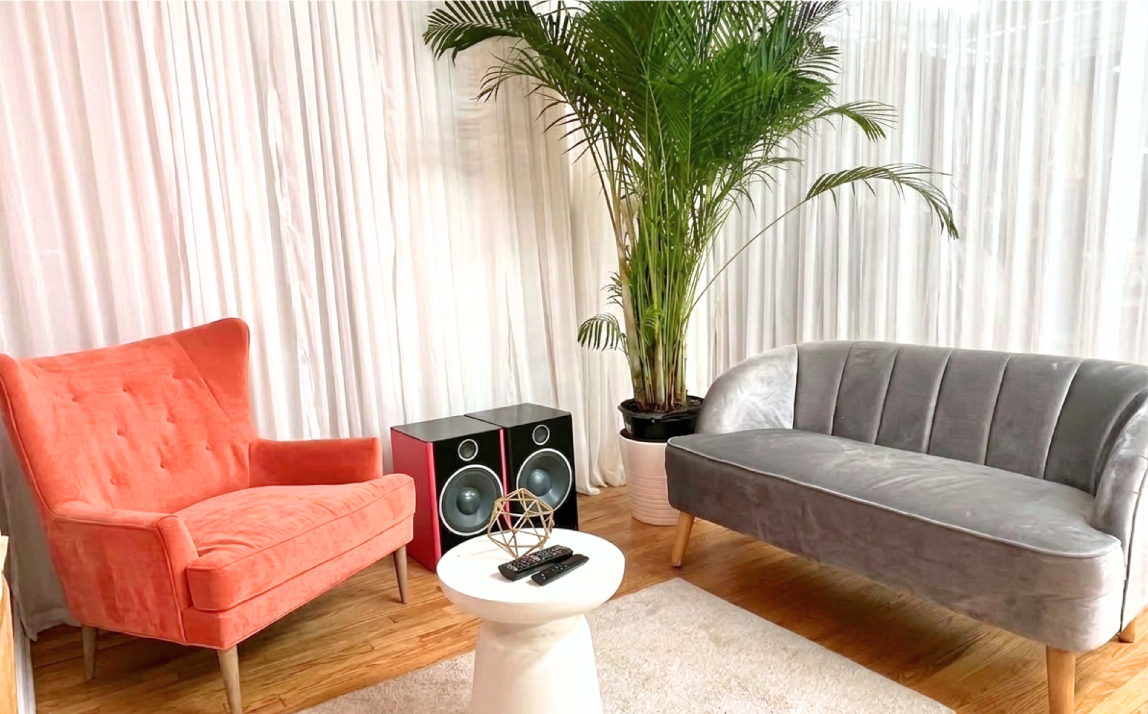 The image shows a cozy living space with a coral armchair, a gray sofa, a small white table, and a speaker, complemented by a tall potted plant in the background.