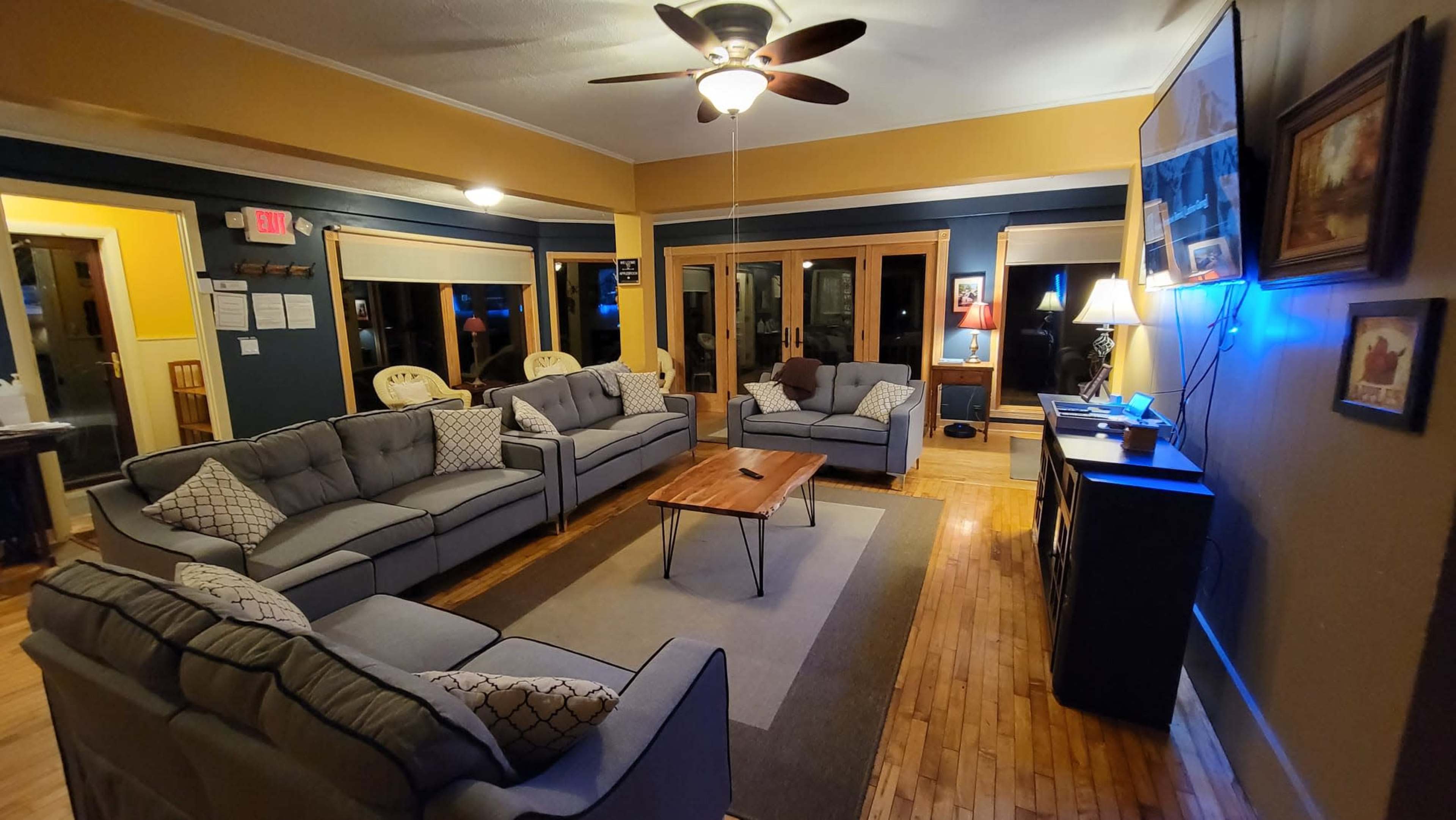 A spacious living room furnished with multiple gray sofas arranged around a wooden coffee table, with large windows providing a view of the outside.