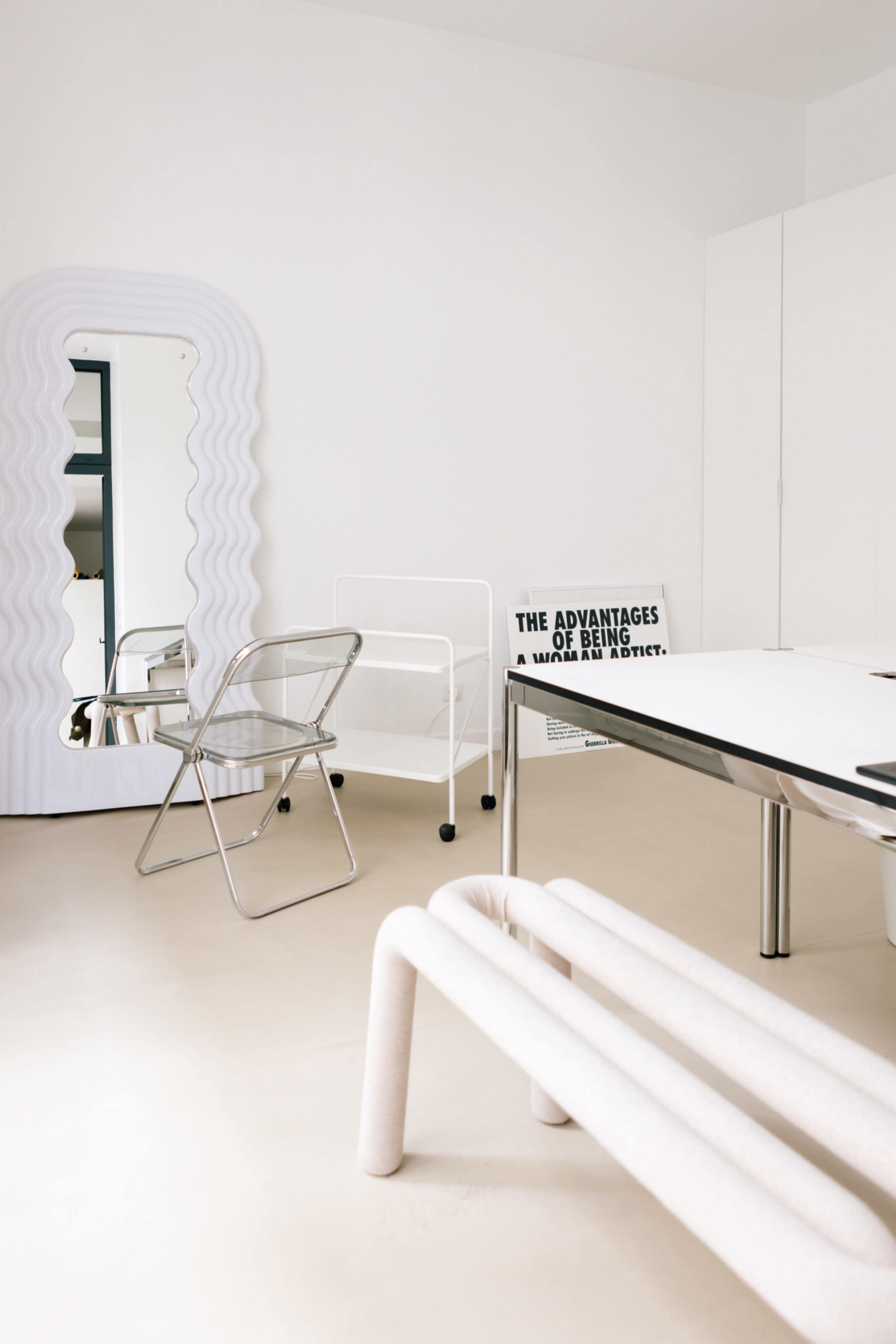 The image shows a minimalist interior space with a wave-patterned mirror, a metallic chair, a rolling cart, and a white table.