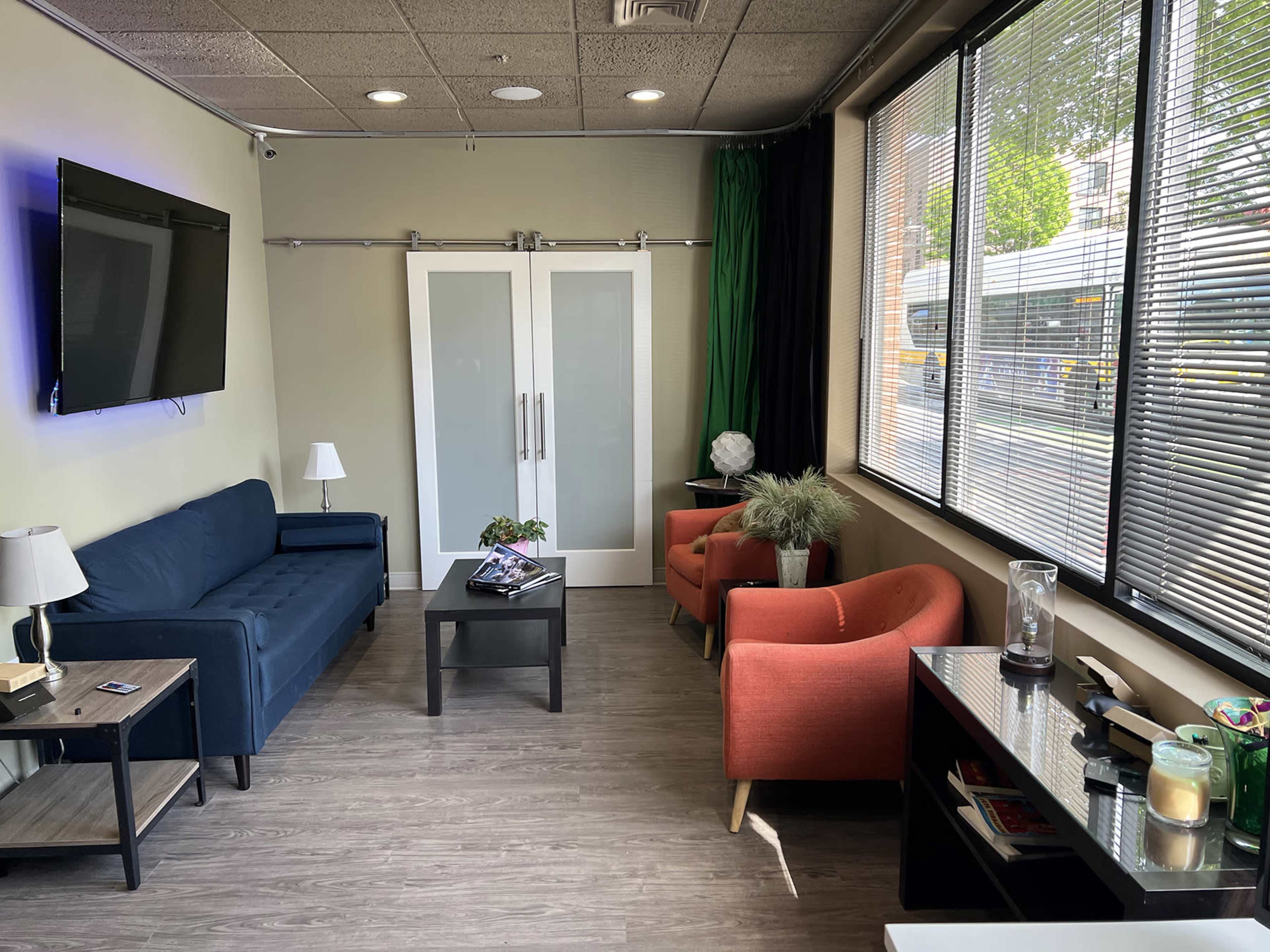 A modern waiting area featuring a blue couch, two orange armchairs, a coffee table, and large windows with blinds, illuminating the space.
