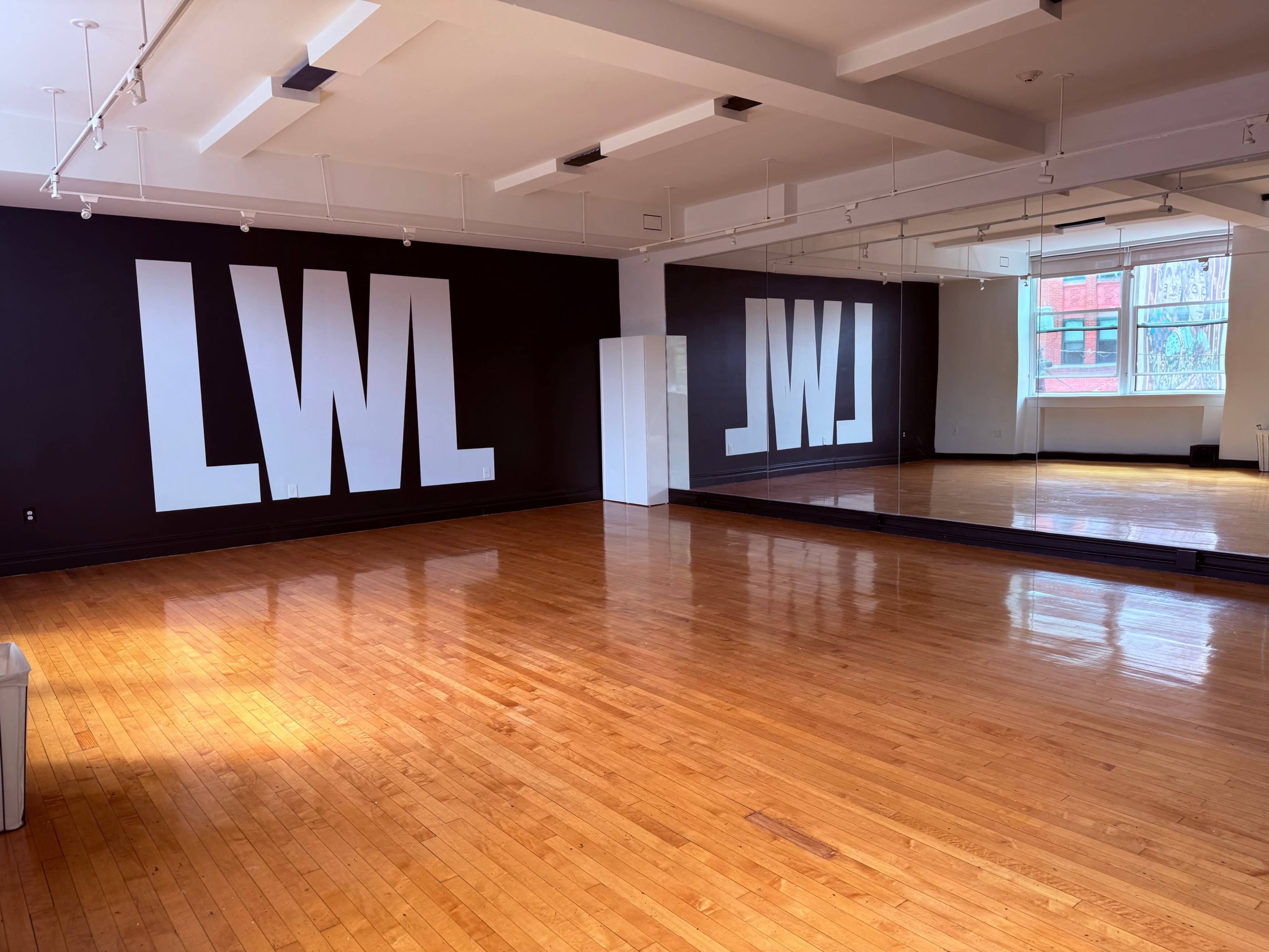The image shows a spacious dance studio with large white letters "LWL" painted on a black wall, and a mirror reflecting the room's wooden floor and windows.