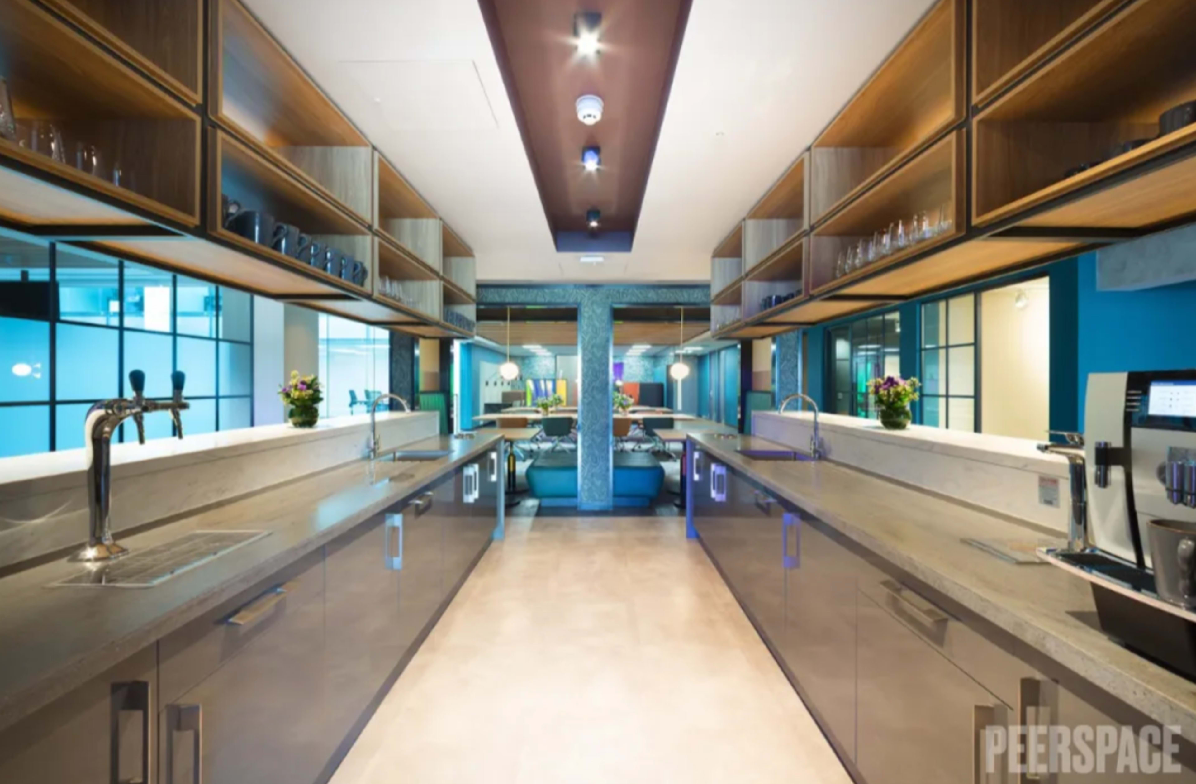 The image shows a modern kitchen with long countertops, shelves above with glassware, and a coffee machine on one side, all set in a bright, contemporary space.